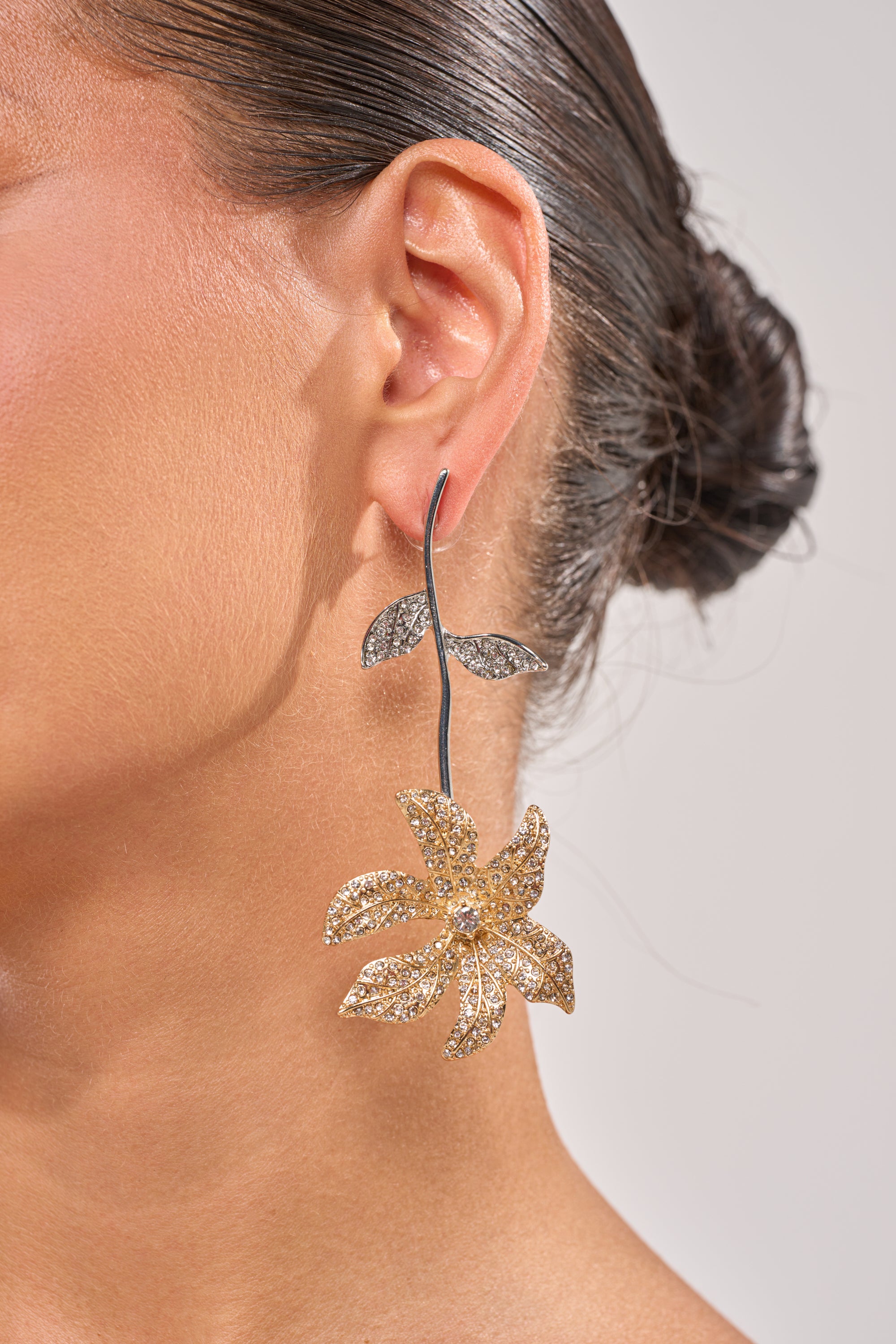 A close-up of a woman's ear adorned with the AT THE GALA EARRING—rhinestone floral drop earrings that sparkle beautifully. Her dark hair is styled in a neat bun, making these perfect wedding guest accessories. 1