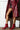 A person dressed in a glossy red coat stands with their legs crossed, showcasing the AZALEA WANG PURNIMA RED BOOTIE featuring stiletto heels, paired with an embroidered black skirt. The faux leather boots stand out against the white wooden backdrop, complementing the chic ensemble.