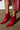 A close-up of someone wearing the AZALEA WANG PURNIMA RED BOOTIE highlights the red, pointed ankle boots featuring distinctive strappy designs and metallic accents. These stiletto heels beautifully enhance a red leather skirt set against a concrete floor and white wall segment.