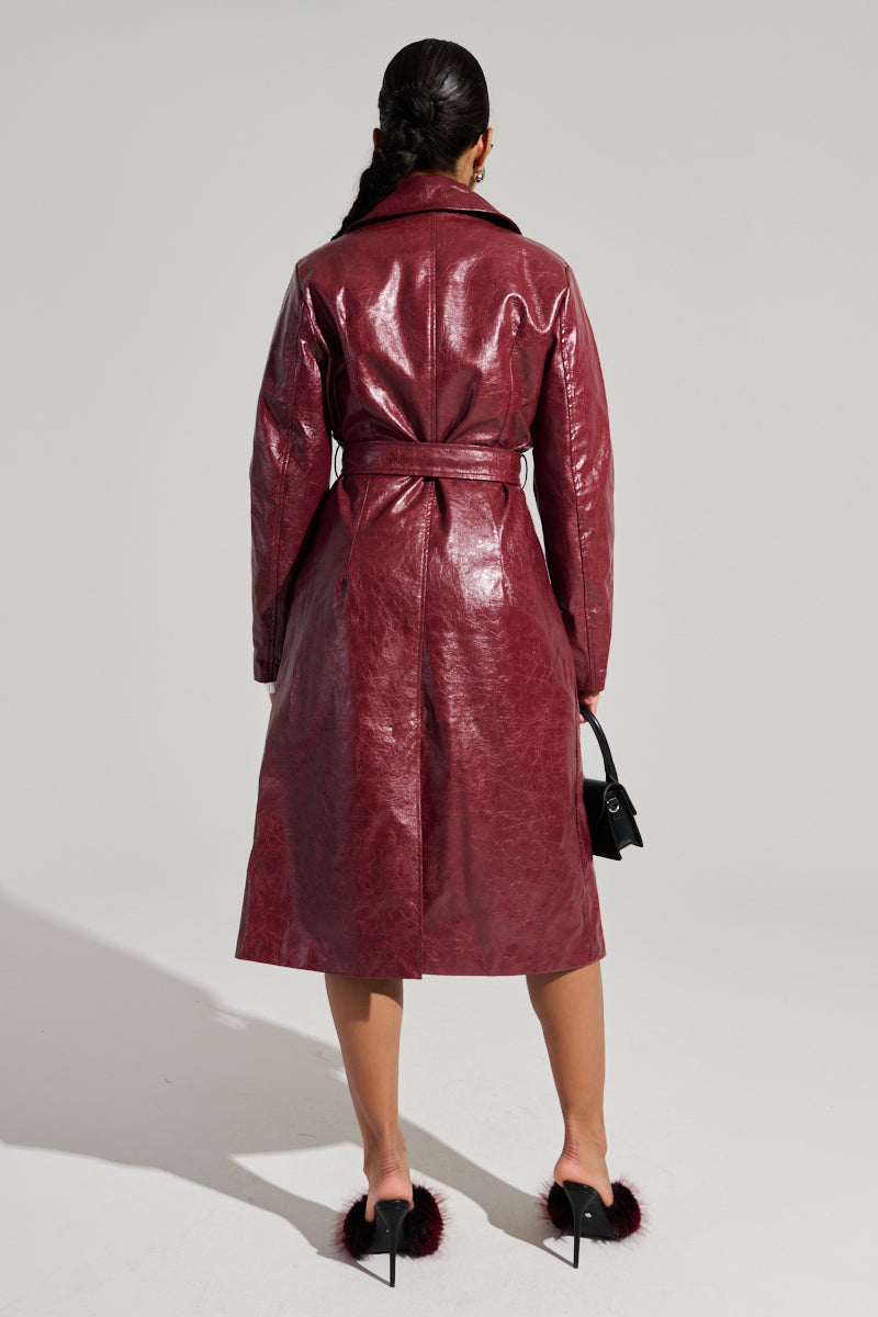 With their back to the camera, a person exudes sophistication in the MISA FAUX LEATHER TRENCH, a shiny burgundy coat complete with a stylish belt. They accessorize with black furry-heeled shoes and hold a small black purse in their left hand against the light gray background.