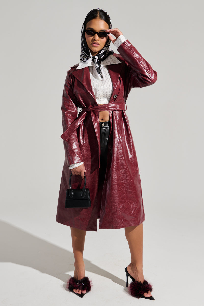 A person stands confidently in a maroon MISA FAUX LEATHER TRENCH coat, black leather pants, and fuzzy high-heeled sandals. They wear a headscarf, sunglasses, and hold a small black handbag. The plain, light-colored background enhances their sophisticated look.