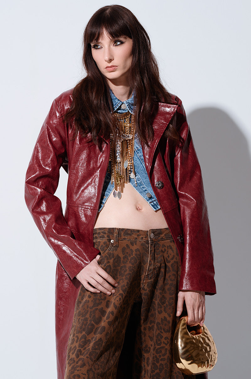 A model poses against a plain background wearing the MISA FAUX LEATHER TRENCH in a shiny red finish over a blue denim shirt and adorned with elaborate gold jewelry. The model's brown hair cascades over their shoulders, while they sport brown leopard-print pants and hold a small gold handbag, completing the sophisticated look.