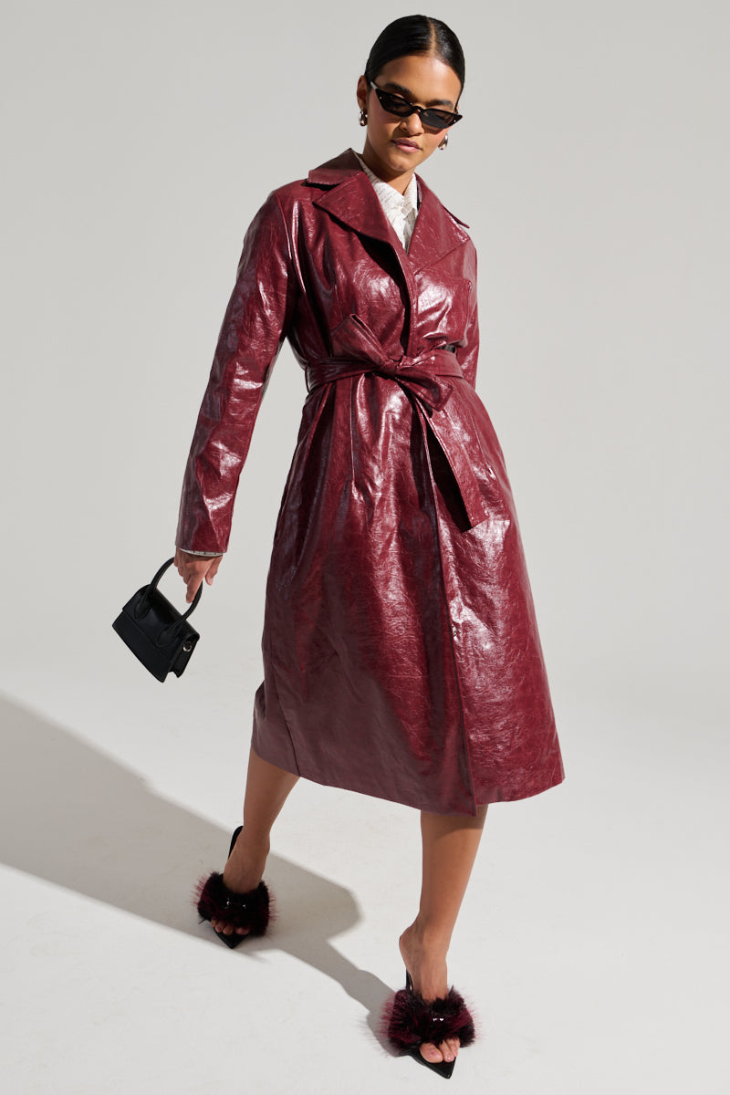 A person models the MISA Faux Leather Trench, a shiny burgundy coat with a tied waist belt. They complement it with black pointed-toe mules featuring fur details, dark sunglasses, and a small black handbag. The simple gray background highlights the sophistication of this non-stretchy outerwear.