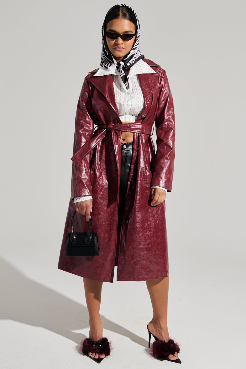 A stylish individual wearing the MISA FAUX LEATHER TRENCH in deep red, paired with black pants and black heels featuring furry accents, accessorized with a black and white headscarf, black sunglasses, and holding a small black handbag, poses confidently against a neutral background for a sophisticated look.
