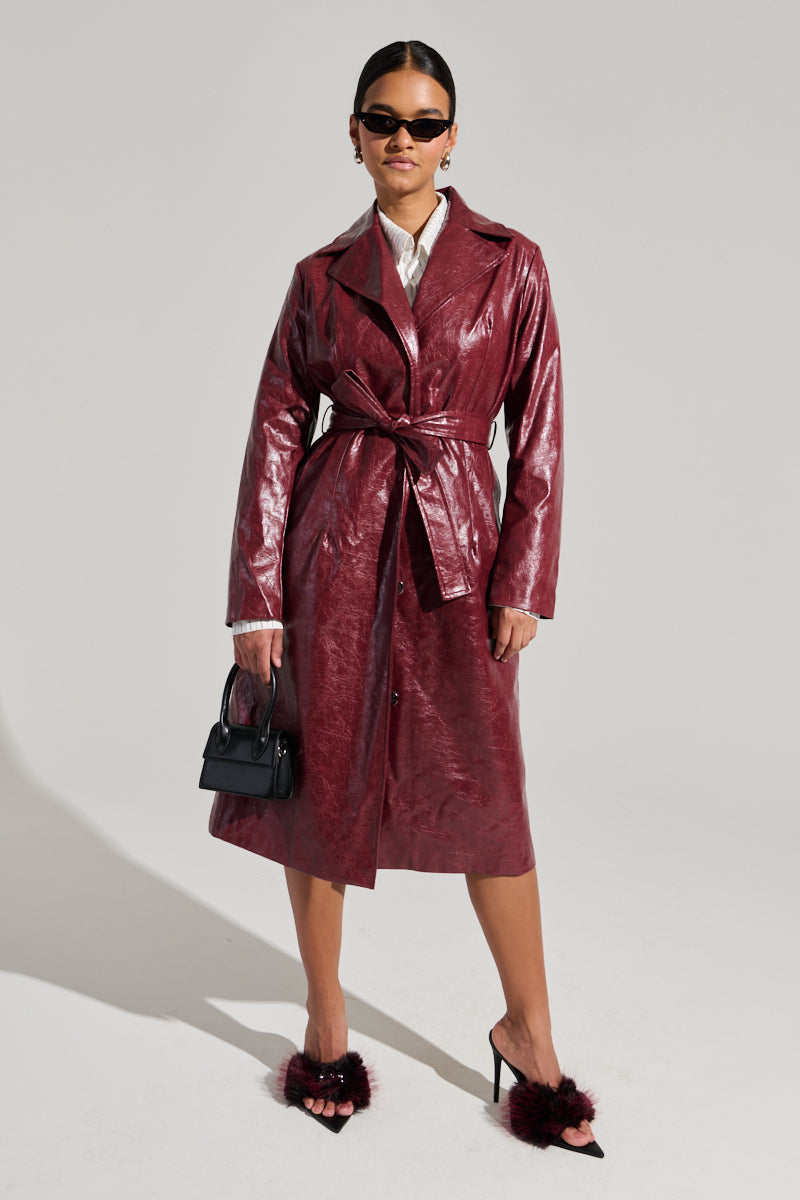 A person confidently poses against a plain background, wearing the MISA FAUX LEATHER TRENCH in shiny, deep-red, tied at the waist. They pair it with black sunglasses, a white blouse, and textured black heels, holding a small black handbag in one hand.