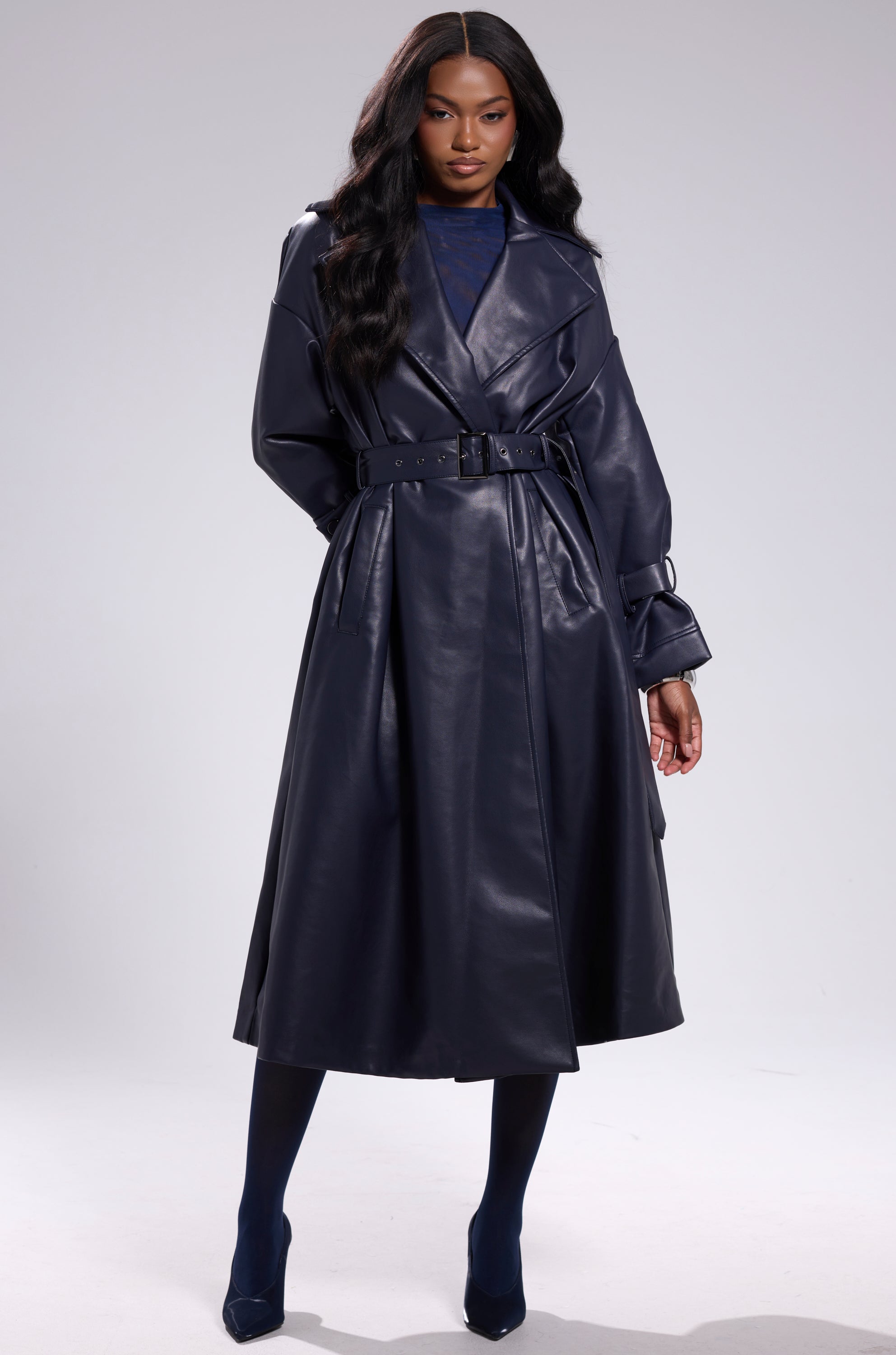 A woman poses confidently against a plain background, wearing the ALINNA FAUX LEATHER TRENCH IN NAVY over a dark top, with matching navy tights and shoes. She has long, wavy hair. 9