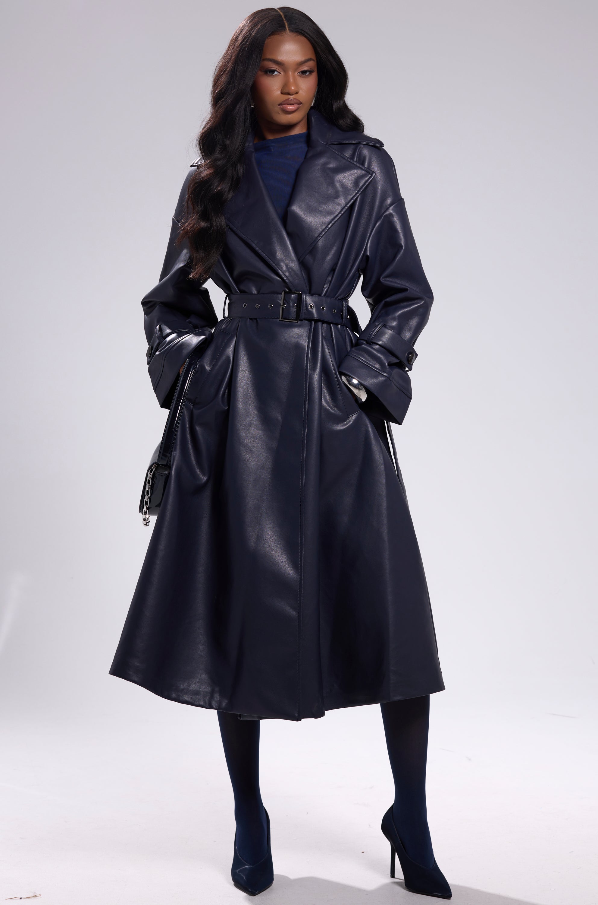 A woman models the ALINNA FAUX LEATHER TRENCH IN NAVY, styled with a matching outfit and heels. She stands confidently with her hands in her pockets, long wavy hair flowing, against a light gray background. 10