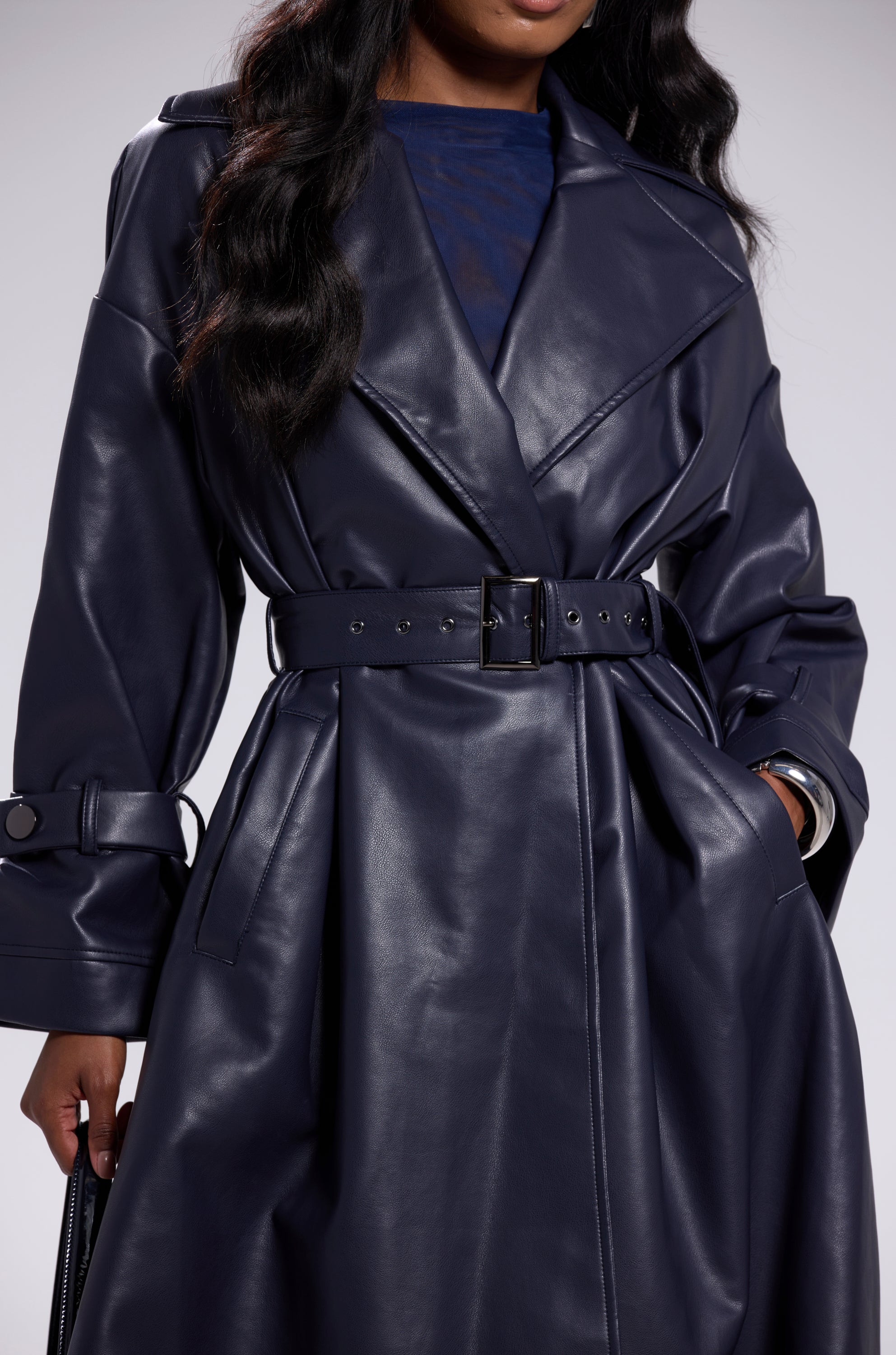 A person wears the ALINNA FAUX LEATHER TRENCH IN NAVY, an oversized coat with wide lapels and long sleeves, while holding a black bag. Their face is partially out of frame. 11