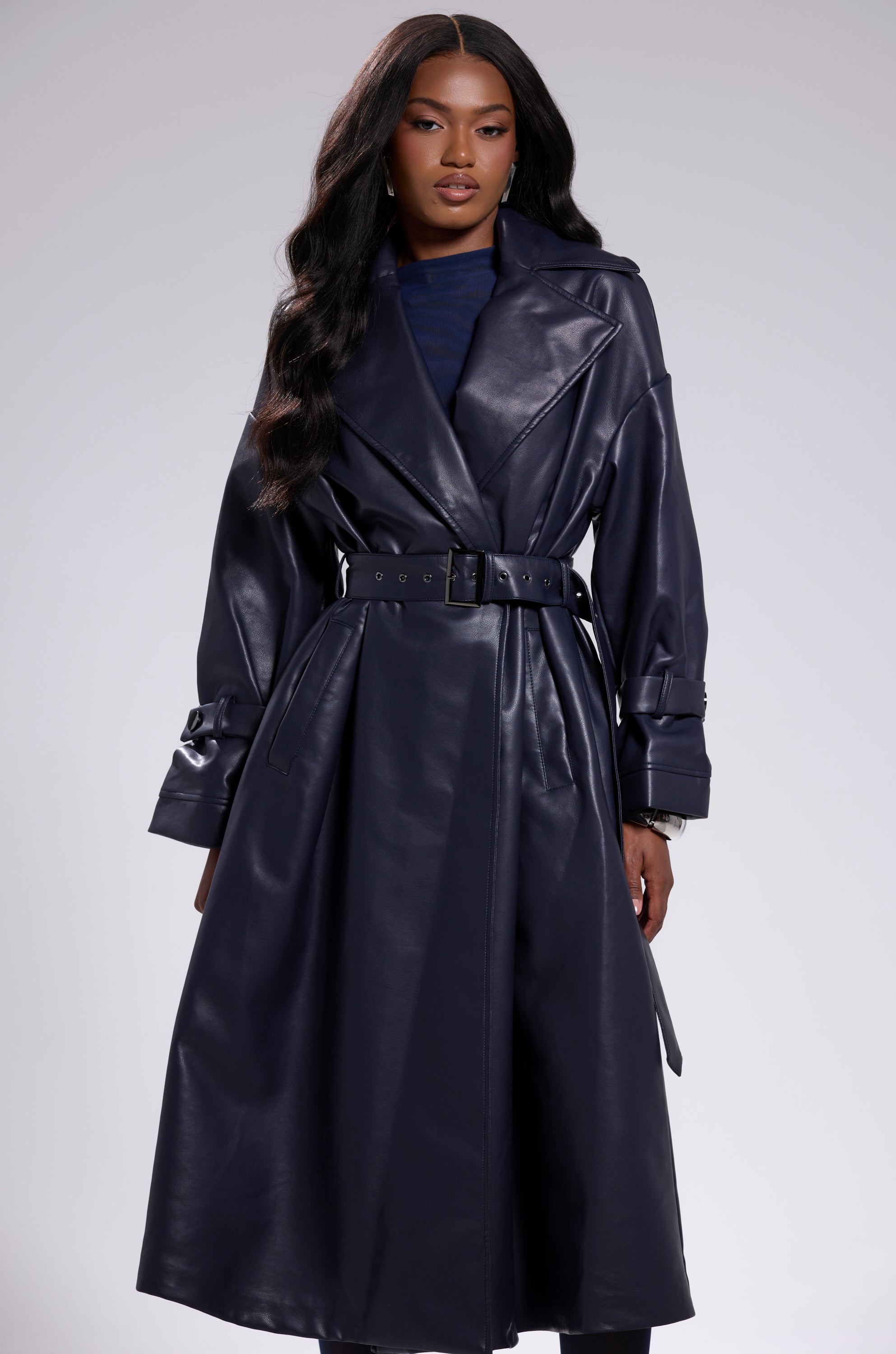 A woman with long wavy hair models the ALINNA FAUX LEATHER TRENCH IN NAVY over a dark top, posing confidently against a plain light gray background. 1