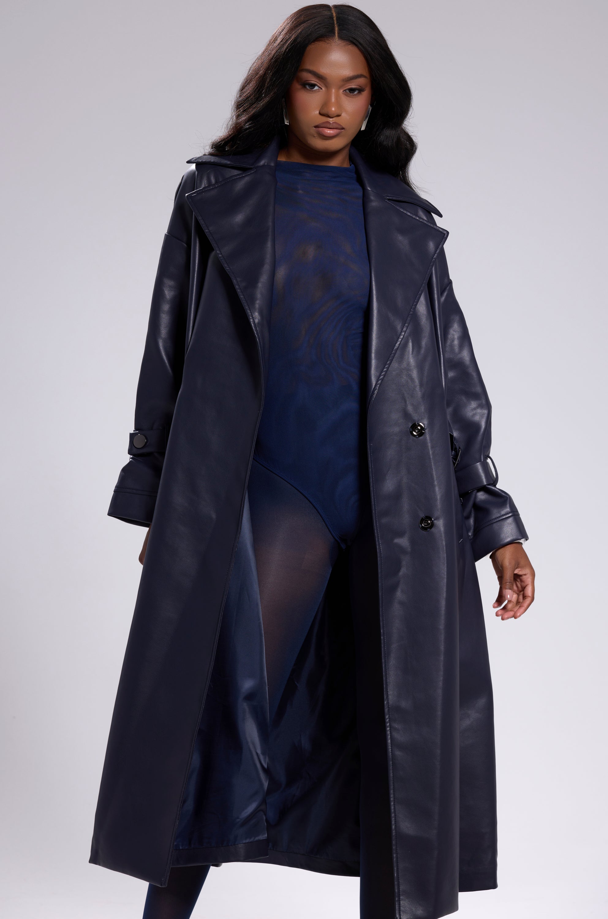 A woman wears the ALINNA FAUX LEATHER TRENCH IN NAVY over a sheer navy bodysuit, posing confidently against a light gray background and highlighting the coat's bold elegance. 3
