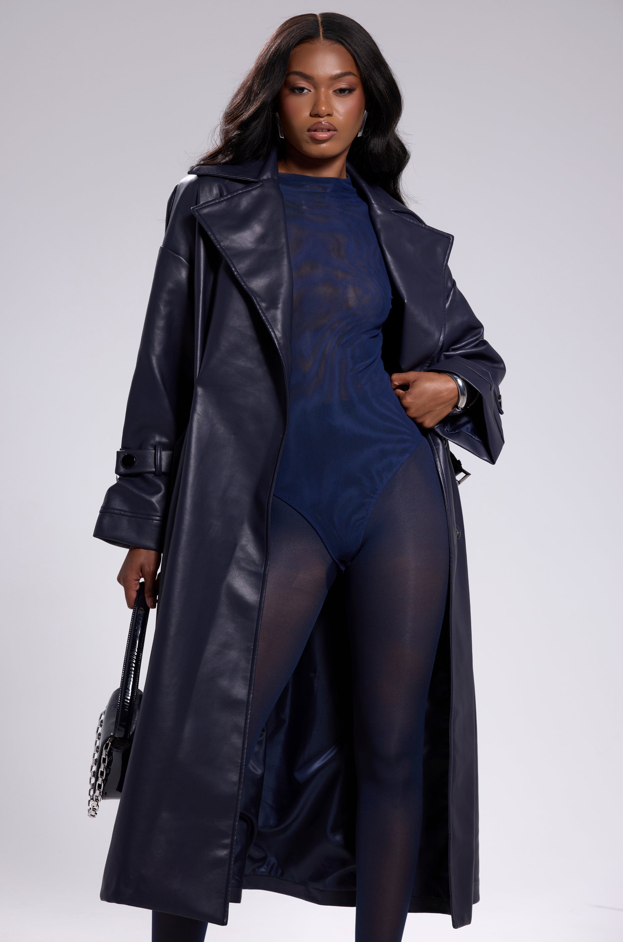 A woman stands confidently in the ALINNA FAUX LEATHER TRENCH IN NAVY over a sheer bodysuit, holding a black handbag with a silver chain. She has long, wavy hair and a neutral expression. 4