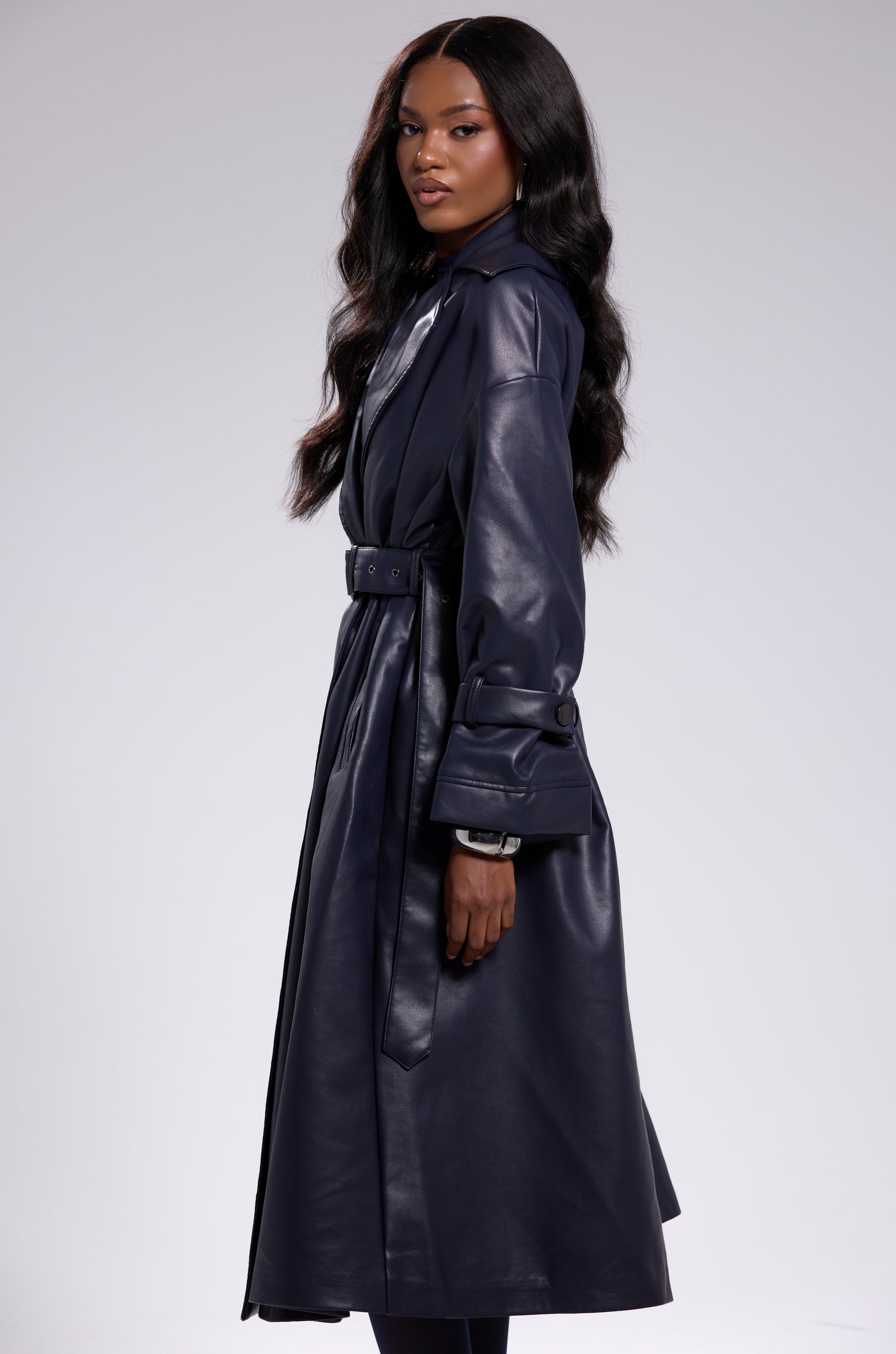 A woman with long wavy hair stands in profile, confidently showcasing the ALINNA FAUX LEATHER TRENCH IN NAVY against a plain light gray background, highlighting its timeless oversized silhouette. 6