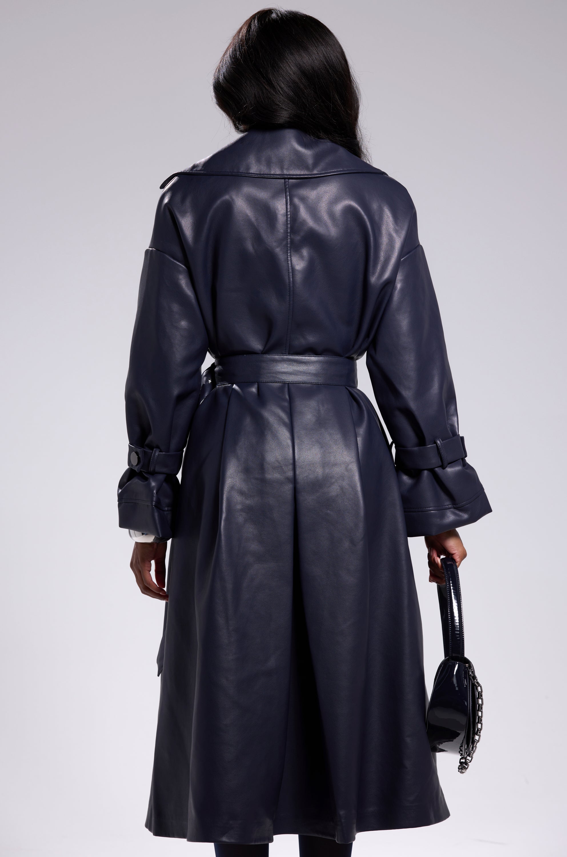 A person with long dark hair, seen from the back, wears the ALINNA FAUX LEATHER TRENCH IN NAVY and holds a small dark purse with a chain strap in their left hand. 7