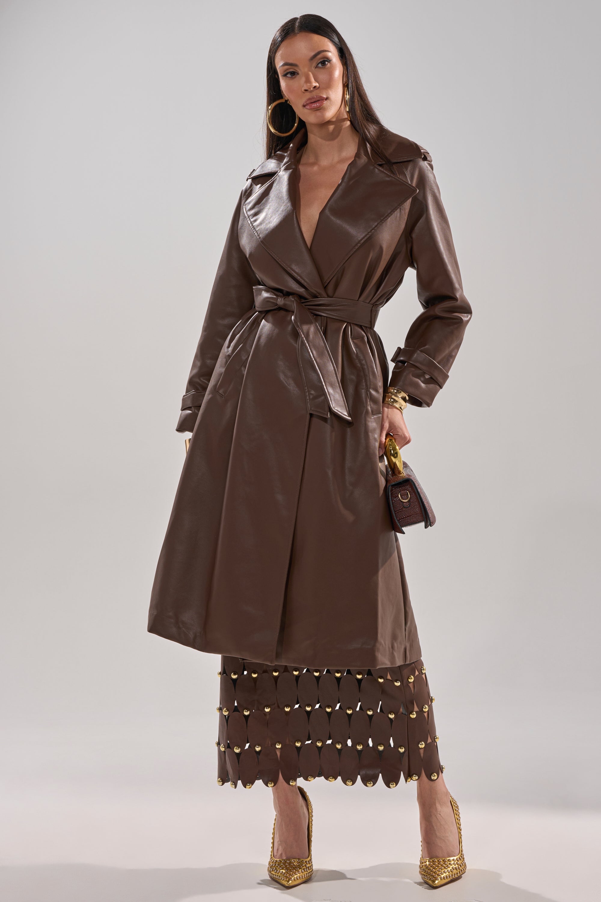 A woman poses confidently in the AKIRA Label ESPRESSO MARTINI FAUX LEATHER TRENCH, gold hoop earrings, a gold and brown cut-out skirt, gold heels, and a small brown handbag against a plain background. 9