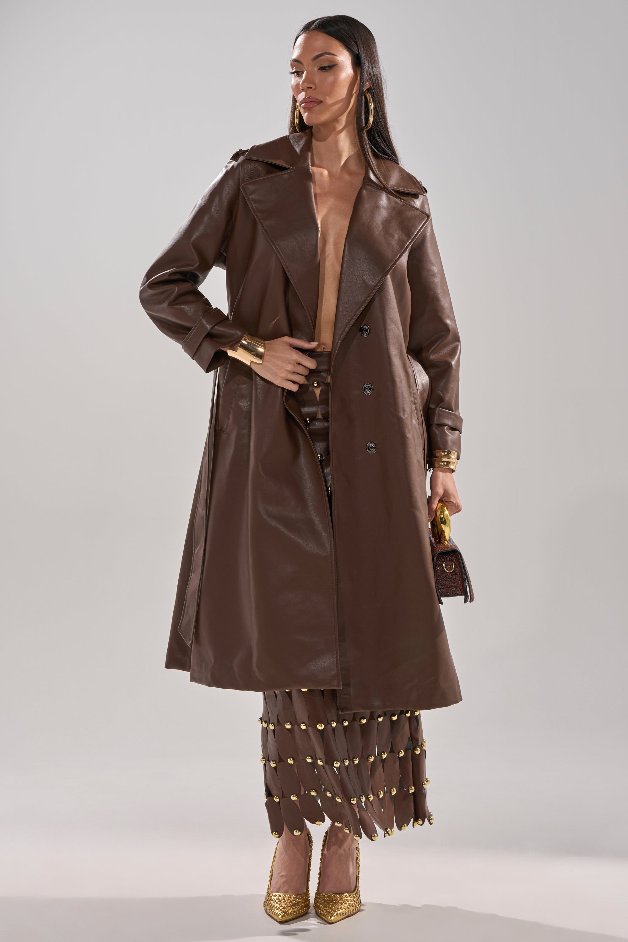 A woman confidently poses in the ESPRESSO MARTINI FAUX LEATHER TRENCH over a gold-embellished brown skirt, gold heels, gold jewelry, and holding a small brown purse against a plain background. 10