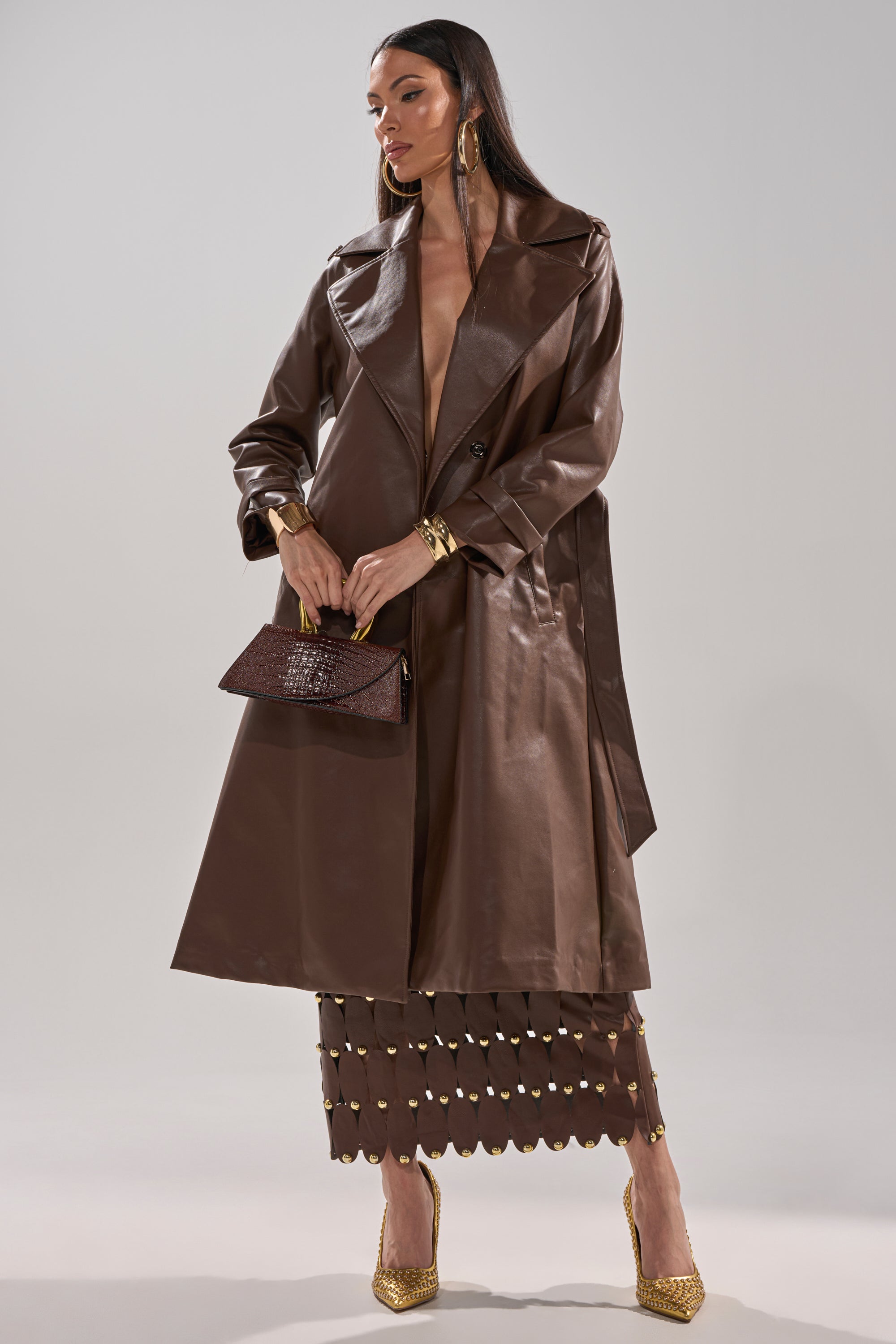 A woman stands confidently against a plain background, wearing the AKIRA Label ESPRESSO MARTINI FAUX LEATHER TRENCH over a brown cutout skirt, with a brown clutch, gold earrings and bracelets, and gold pointed heels. 11