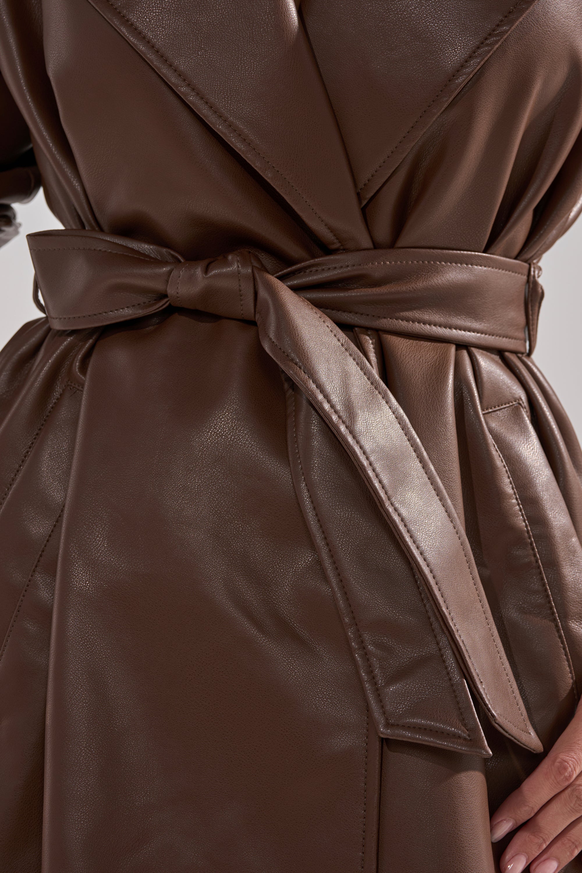 Close-up of a person wearing the ESPRESSO MARTINI FAUX LEATHER TRENCH in brown, featuring a wide belt tied in a bow at the waist and visible stitching details. 12