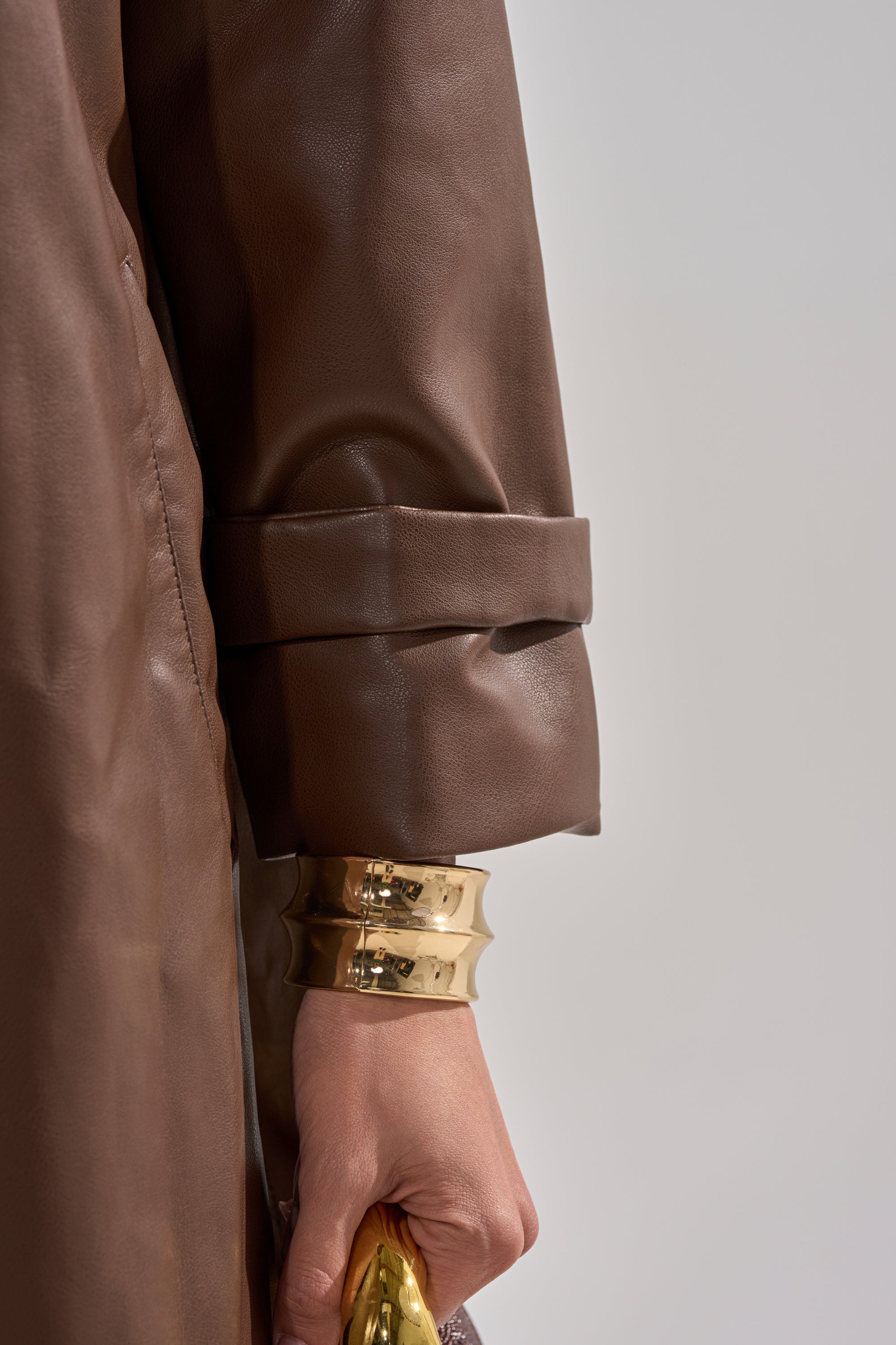 A close-up of a person’s arm wearing the ESPRESSO MARTINI FAUX LEATHER TRENCH with a cuffed sleeve, featuring two gold bracelets and holding a gold object against a plain, light background. 13
