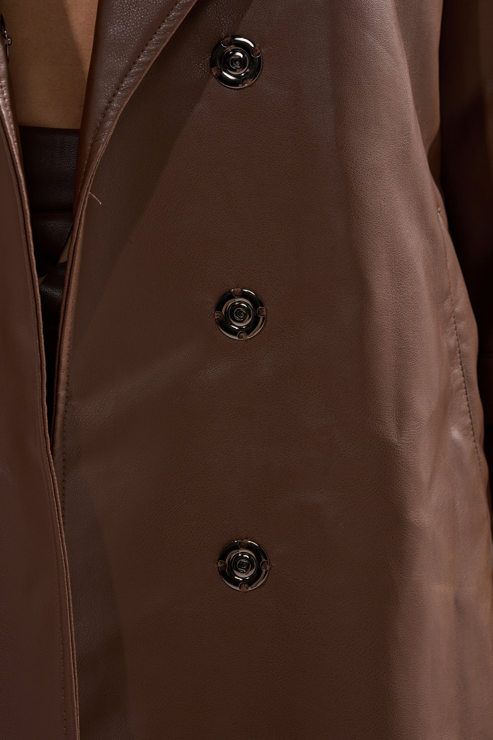 A close-up of the ESPRESSO MARTINI FAUX LEATHER TRENCH, a brown faux leather longline coat with three shiny black buttons, worn slightly open to reveal a hint of a brown garment underneath. 14