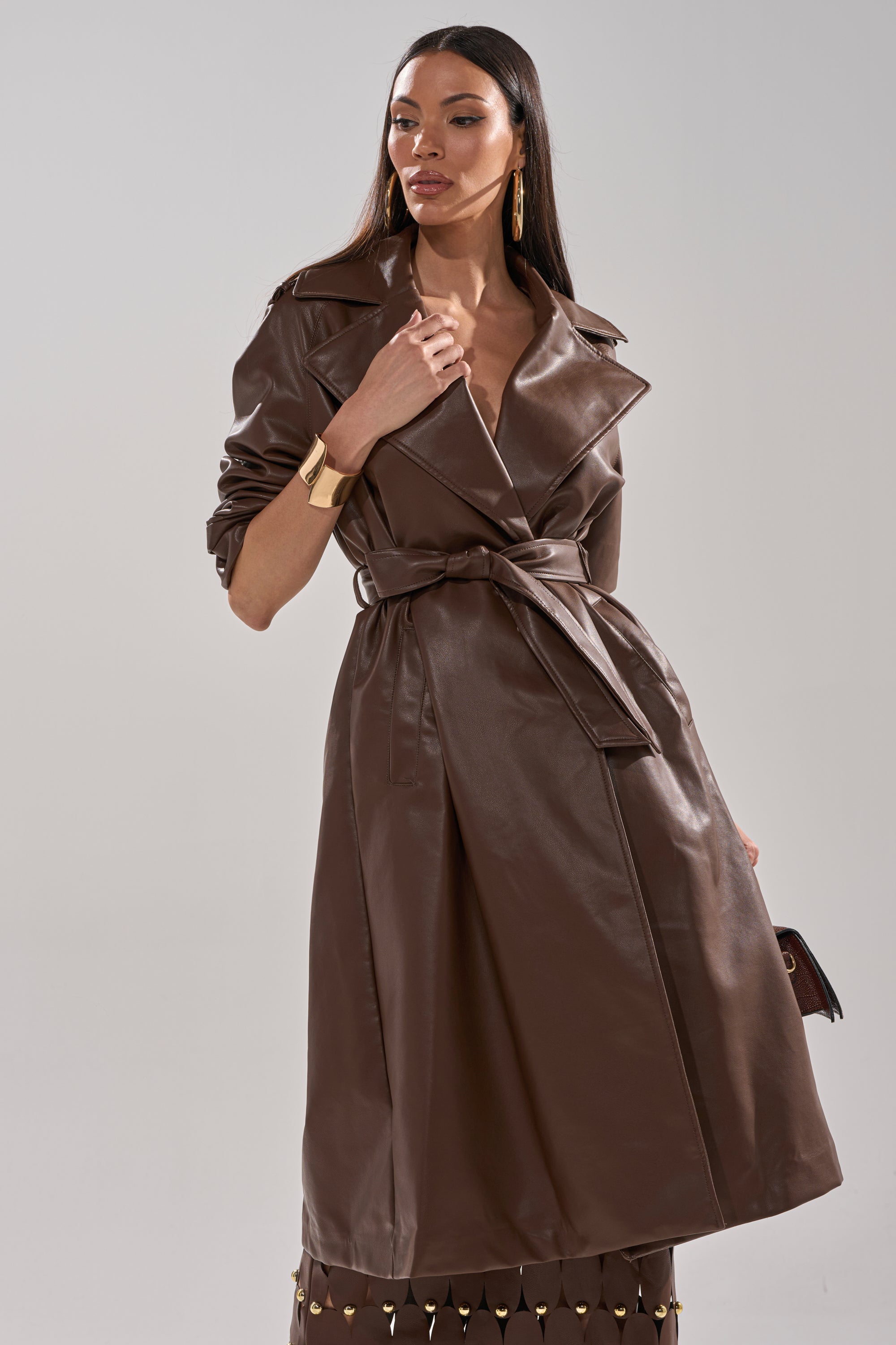 A woman poses confidently against a plain light background, wearing the ESPRESSO MARTINI FAUX LEATHER TRENCH with a waist belt, paired with gold hoop earrings and a gold bracelet. 1