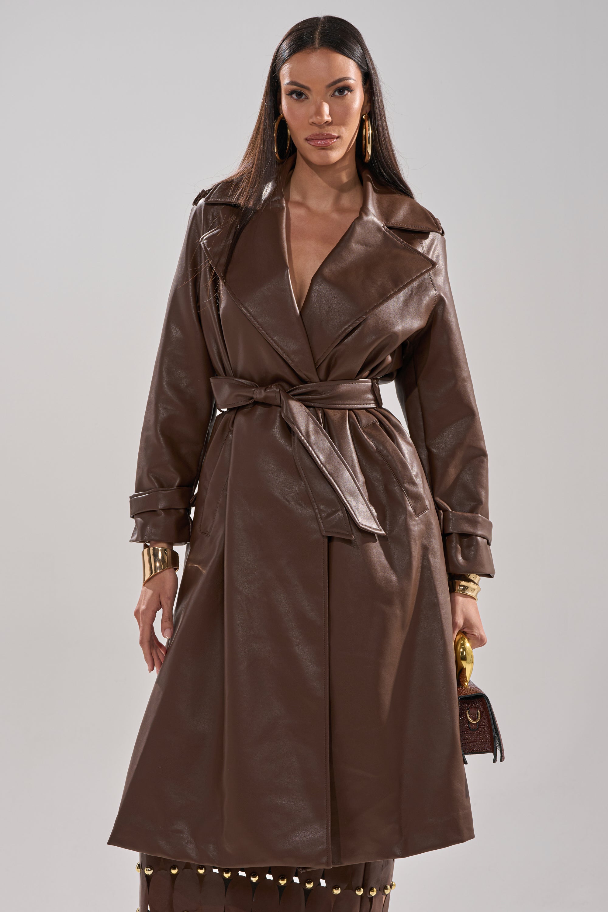 A woman stands confidently against a plain light background, wearing the ESPRESSO MARTINI FAUX LEATHER TRENCH, gold hoop earrings, gold bracelets, and holding a small brown handbag. 2