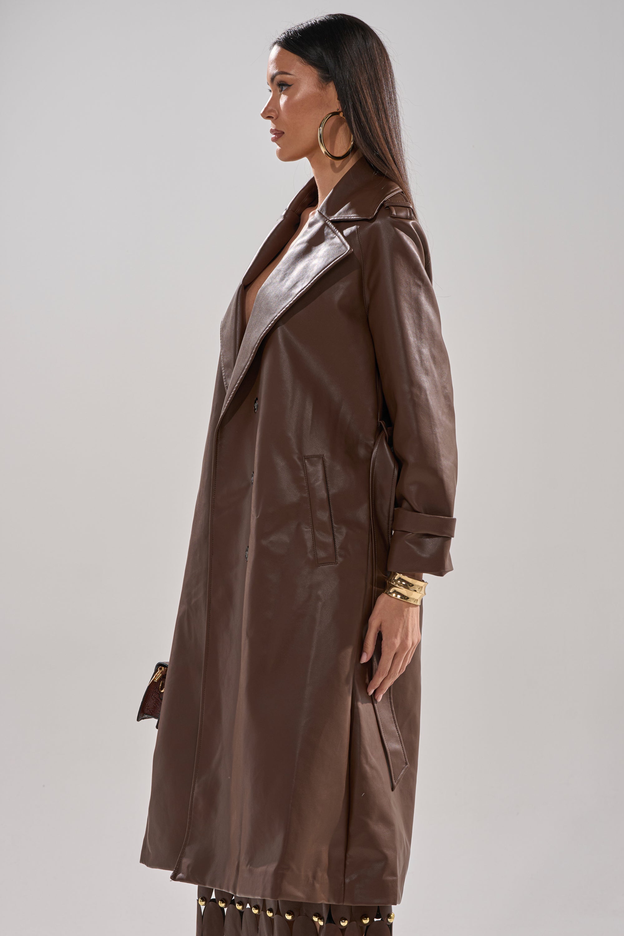 A woman stands in profile wearing the ESPRESSO MARTINI FAUX LEATHER TRENCH, styled with gold hoop earrings and bracelets, against a plain light background. 4