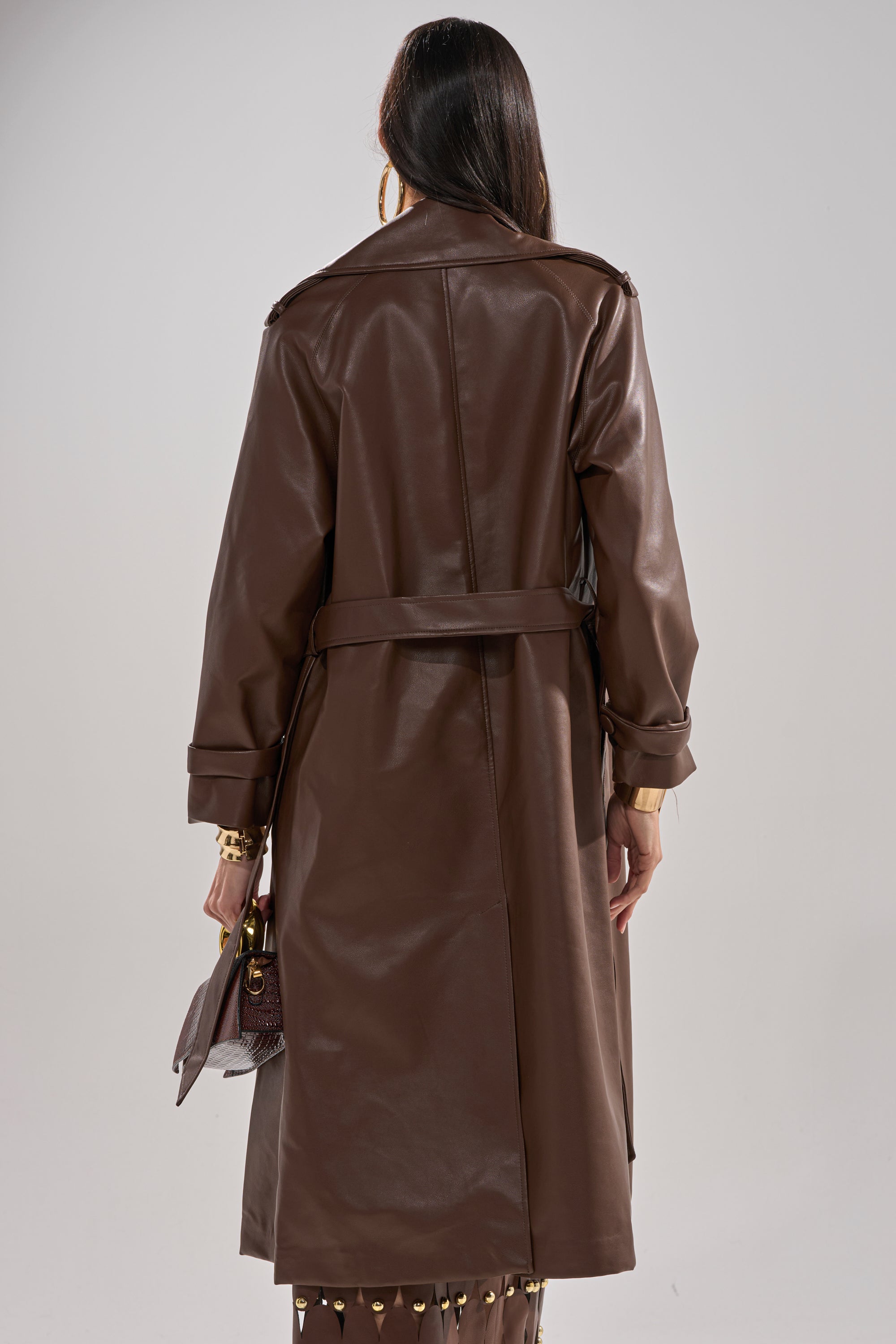 A woman faces away, wearing the ESPRESSO MARTINI FAUX LEATHER TRENCH with a belt, holding a small textured handbag. Her straight dark hair is loose, accessorized with gold hoop earrings and bracelets. 5