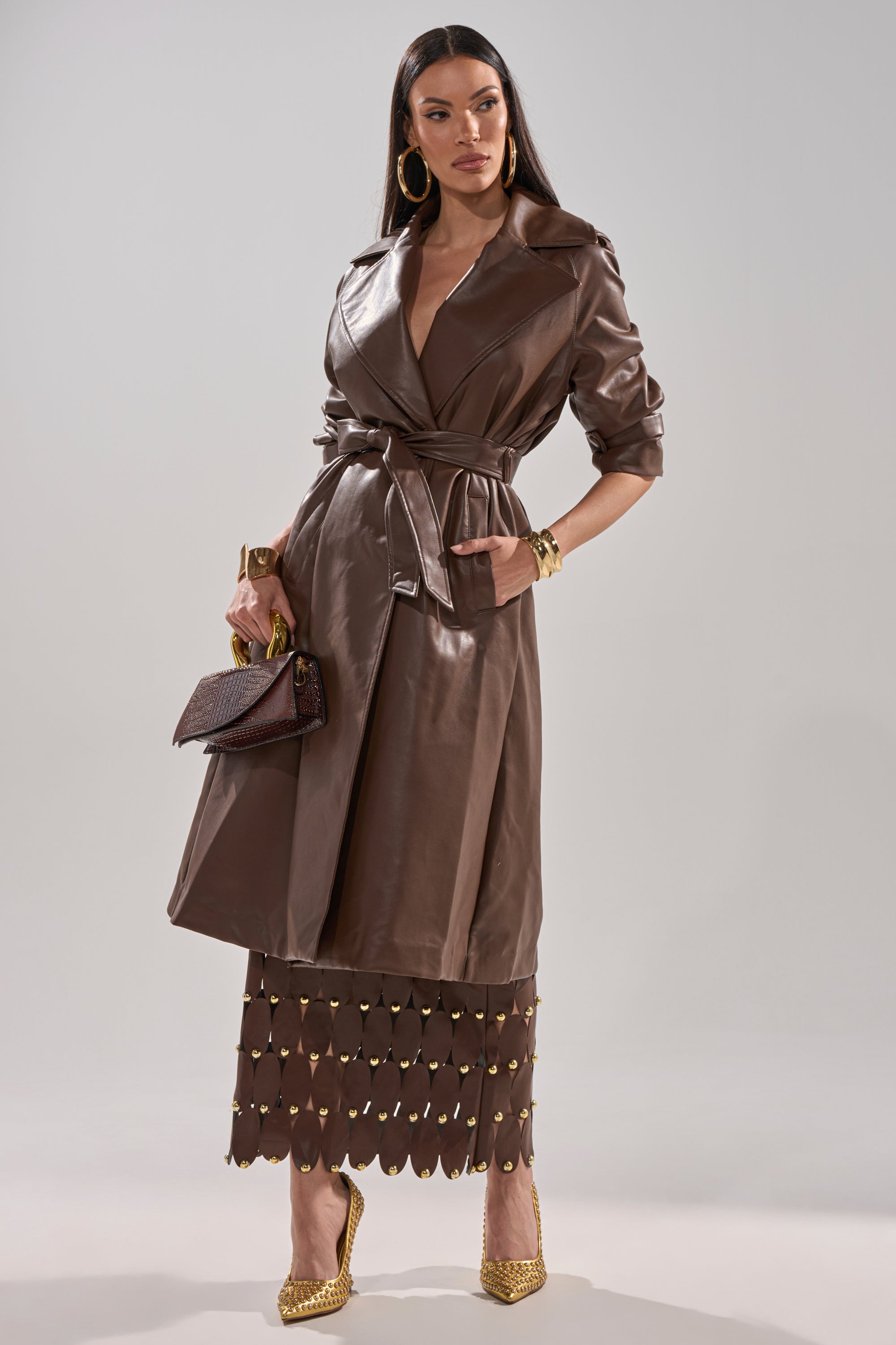 A woman models the ESPRESSO MARTINI FAUX LEATHER TRENCH over a brown skirt with gold heels, hoop earrings, gold bracelets, and a brown textured handbag, posing against a plain background. 6
