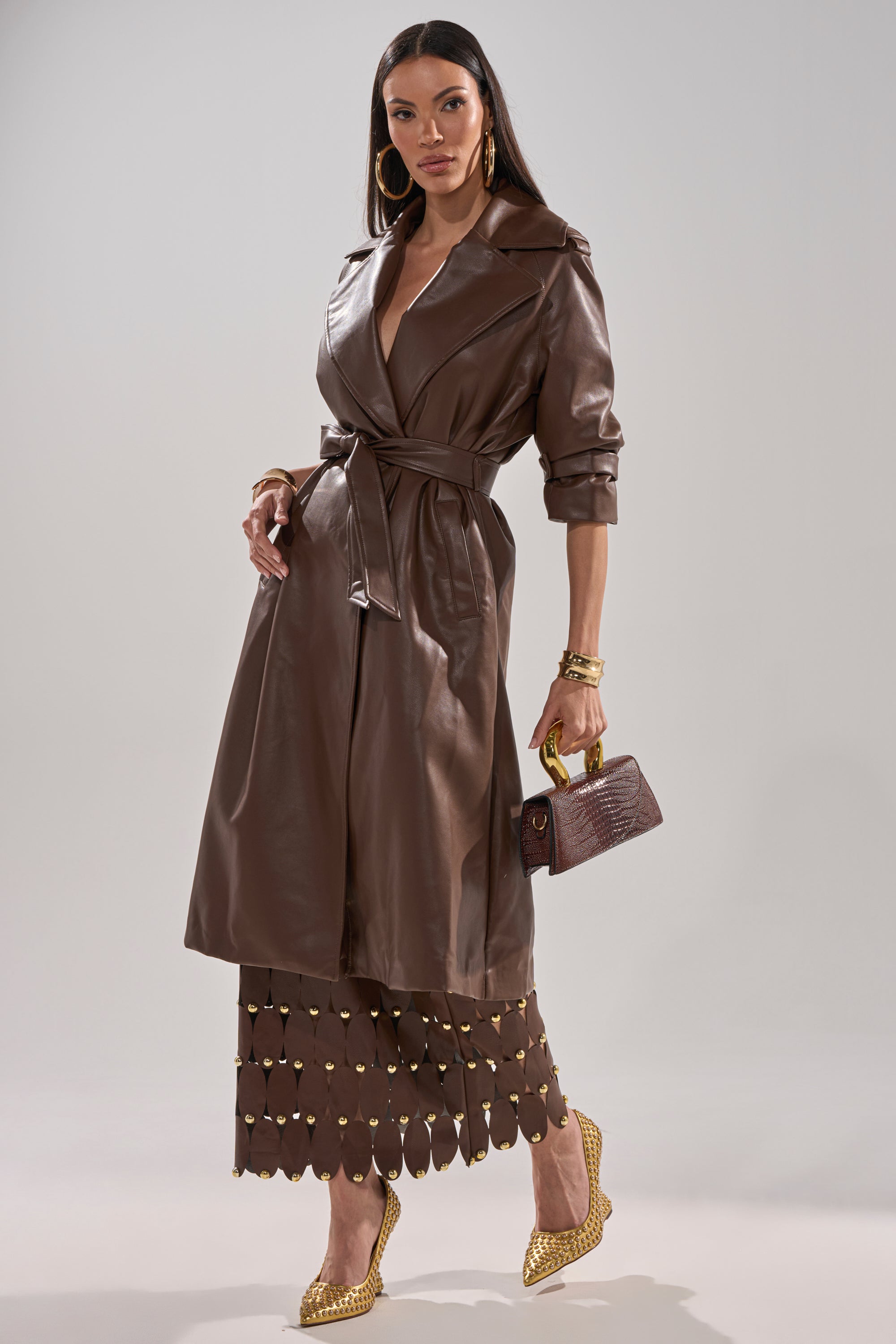A woman wears the ESPRESSO MARTINI FAUX LEATHER TRENCH in a longline silhouette, paired with a brown skirt, gold accents, gold shoes, hoop earrings, bracelets, and a small brown handbag, posing against a neutral background. 7