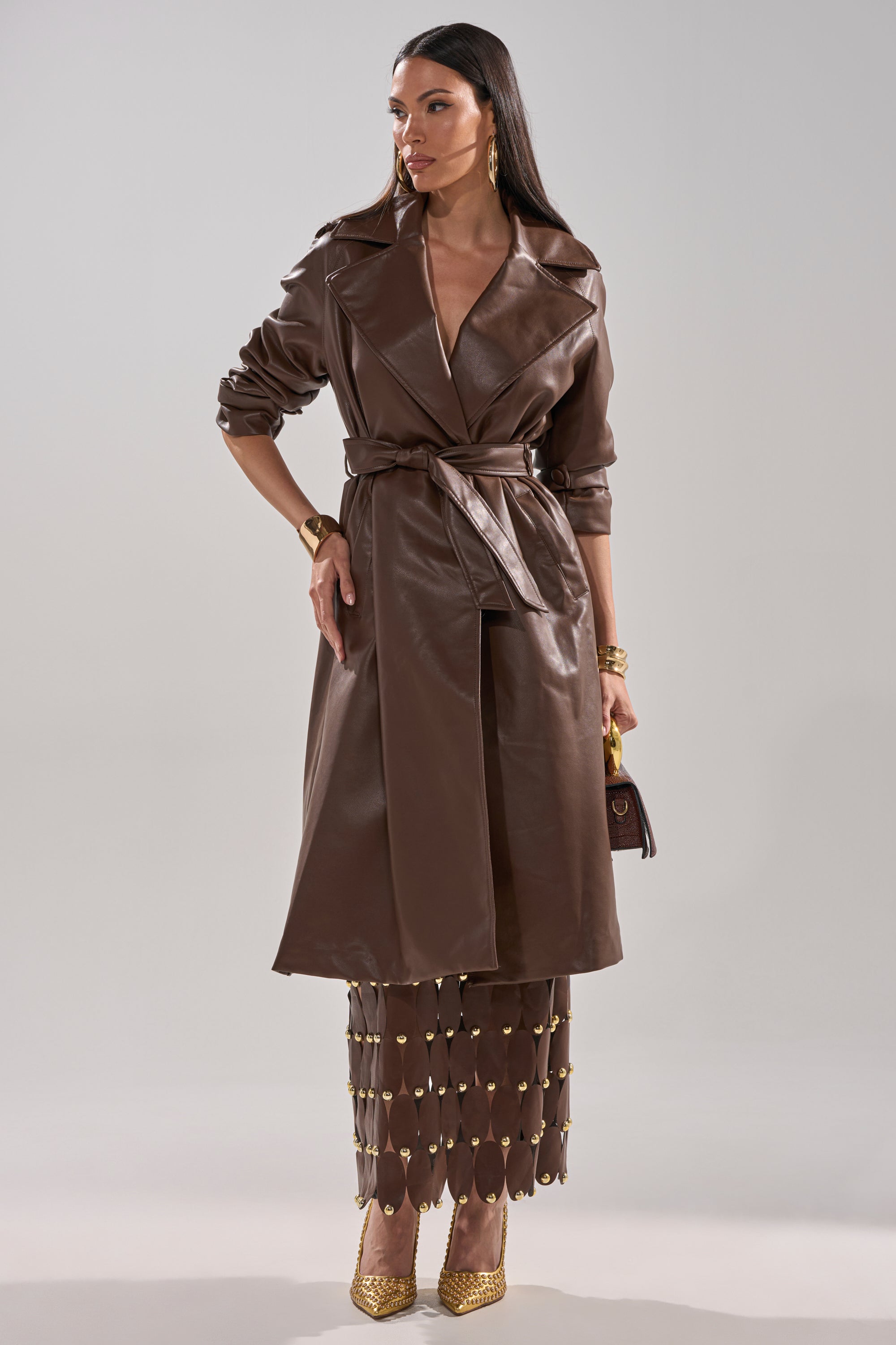 A woman wears the ESPRESSO MARTINI FAUX LEATHER TRENCH tied at the waist, paired with a long brown skirt with gold accents, gold heels, gold jewelry, and a small brown handbag against a plain light gray background. 8