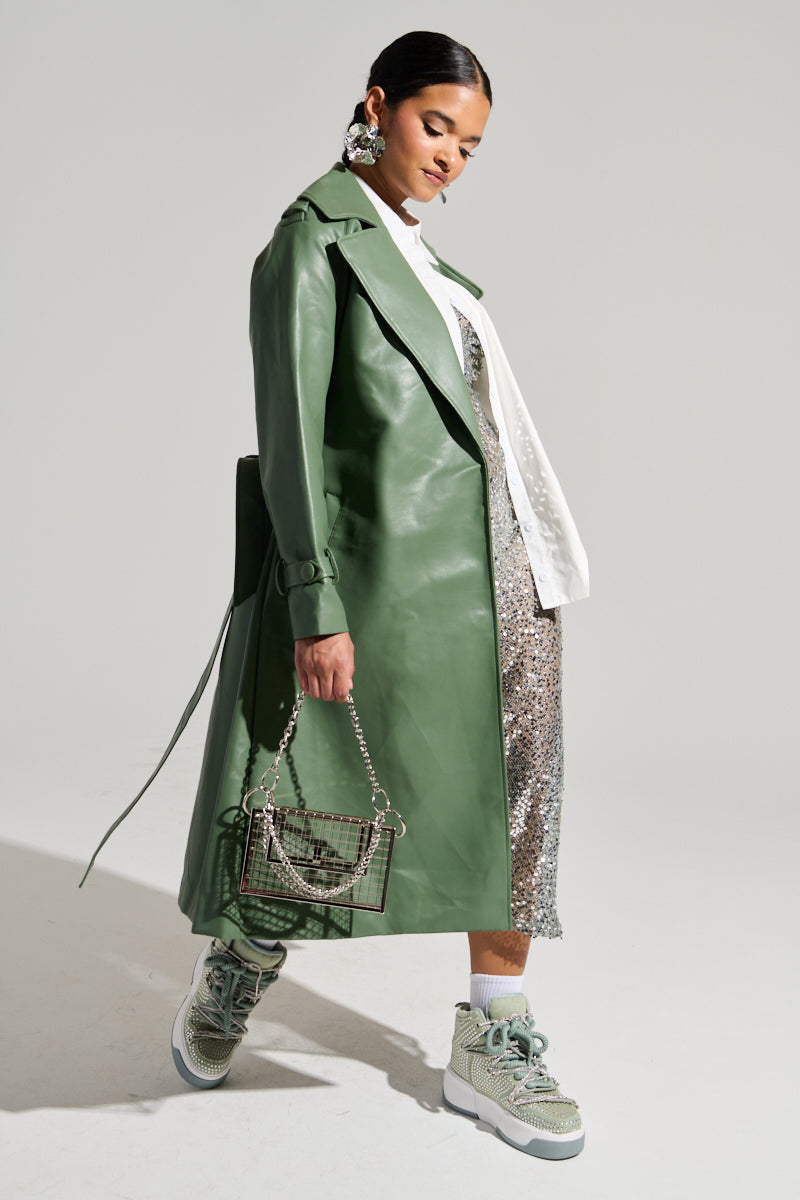 A fashion-forward individual poses in the DIRTY MARTINI FAUX LEATHER TRENCH with an oversized notch lapel collar, draped over a silver sequined dress. They hold a small, stylish handbag and wear chunky, light green sneakers. Their hair is styled sleekly, and they have bold, decorative earrings.