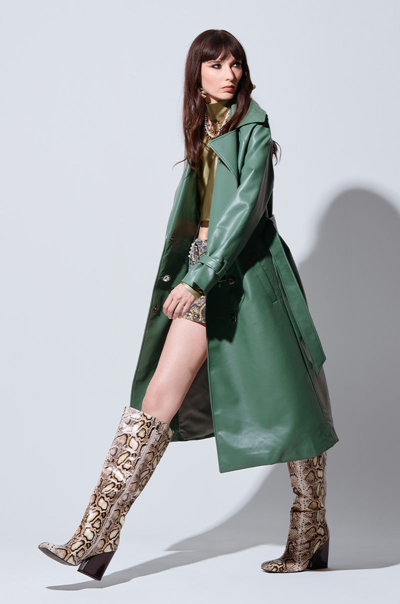 A woman poses in the DIRTY MARTINI FAUX LEATHER TRENCH, highlighting its oversized notch lapel collar and self-tie waist belt closure. She complements it with a turtleneck top, a high-waisted printed mini skirt, and knee-high snakeskin boots. Her long, wavy brown hair cascades against the plain, light grey background.