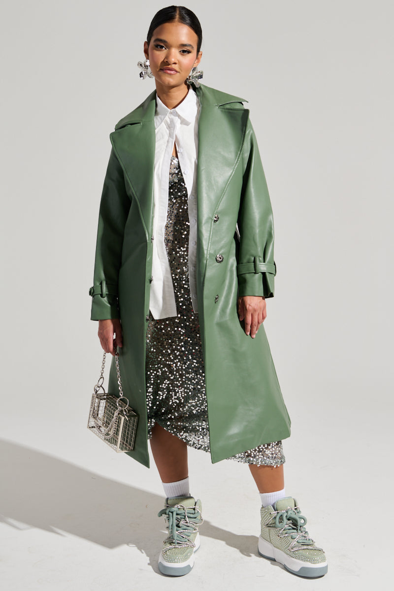 A person stands against a plain background wearing a green DIRTY MARTINI FAUX LEATHER TRENCH, featuring an oversized notch lapel collar, over a white shirt and a sparkly silver dress. They hold a small handbag and wear light-colored sneakers. Their hair is pulled back, and they're adorned with large earrings.