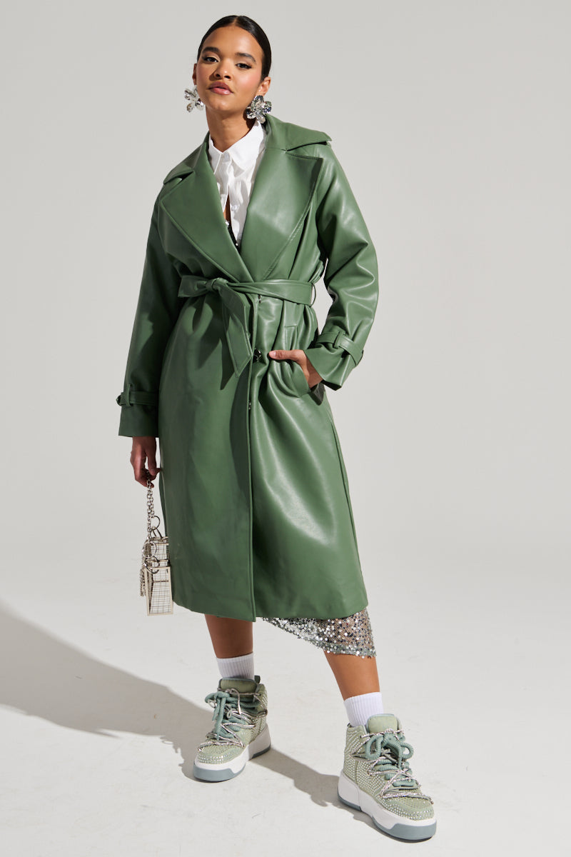 Against a simple backdrop, a woman confidently models the green Dirty Martini Faux Leather Trench, featuring an oversized notch lapel collar. She pairs it with a white blouse and a glittering silver skirt, accessorizing her look with large earrings, a small handbag, and gray sneakers. With one hand in her pocket, she wears an assured expression.