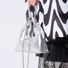 A person wearing a black and white wavy patterned coat and snake print pants holds the chic TOO TRENDY PURSE, a faux silver metallic handbag with a chain strap, against a plain white background.