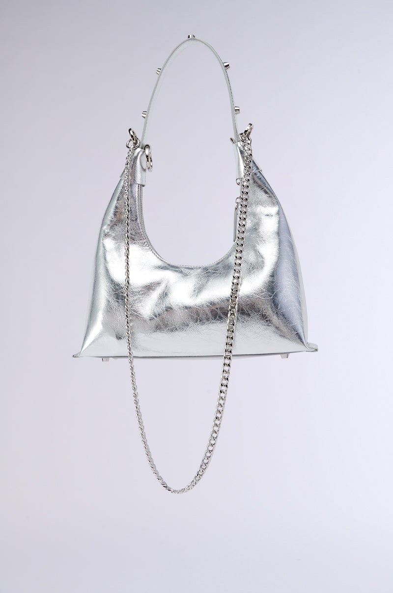 The TOO TRENDY PURSE from AKIRA Label in faux silver metallic showcases a sleek, minimalist design with a curved shape and a long chain strap. A short handle adorned with small metal studs adds a modern, edgy touch against the plain light gray background. 3