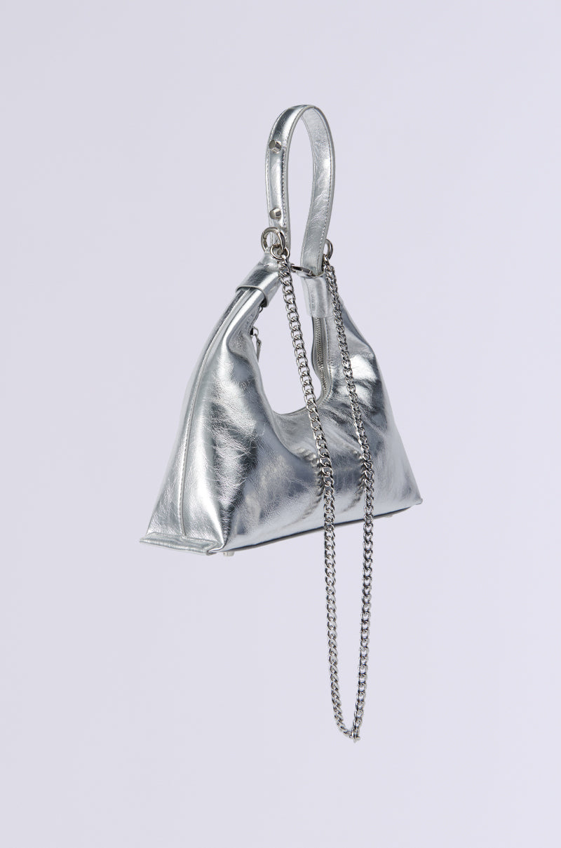 The TOO TRENDY PURSE is a faux silver metallic triangular handbag with a glossy finish, featuring a chic short handle and an elegant long chain strap as an accessory. Its modern and minimalist design is showcased against a plain white background.