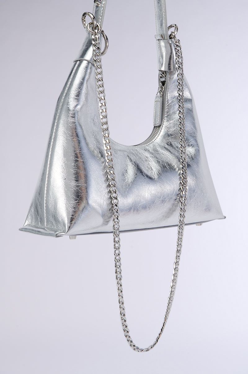 The TOO TRENDY PURSE, a sleek and faux silver metallic handbag with a triangular shape, hangs against a plain background. It features a short handle and a stud-embellished shoulder strap. Its exterior boasts a slight reflective sheen, adding a modern and stylish touch. 5
