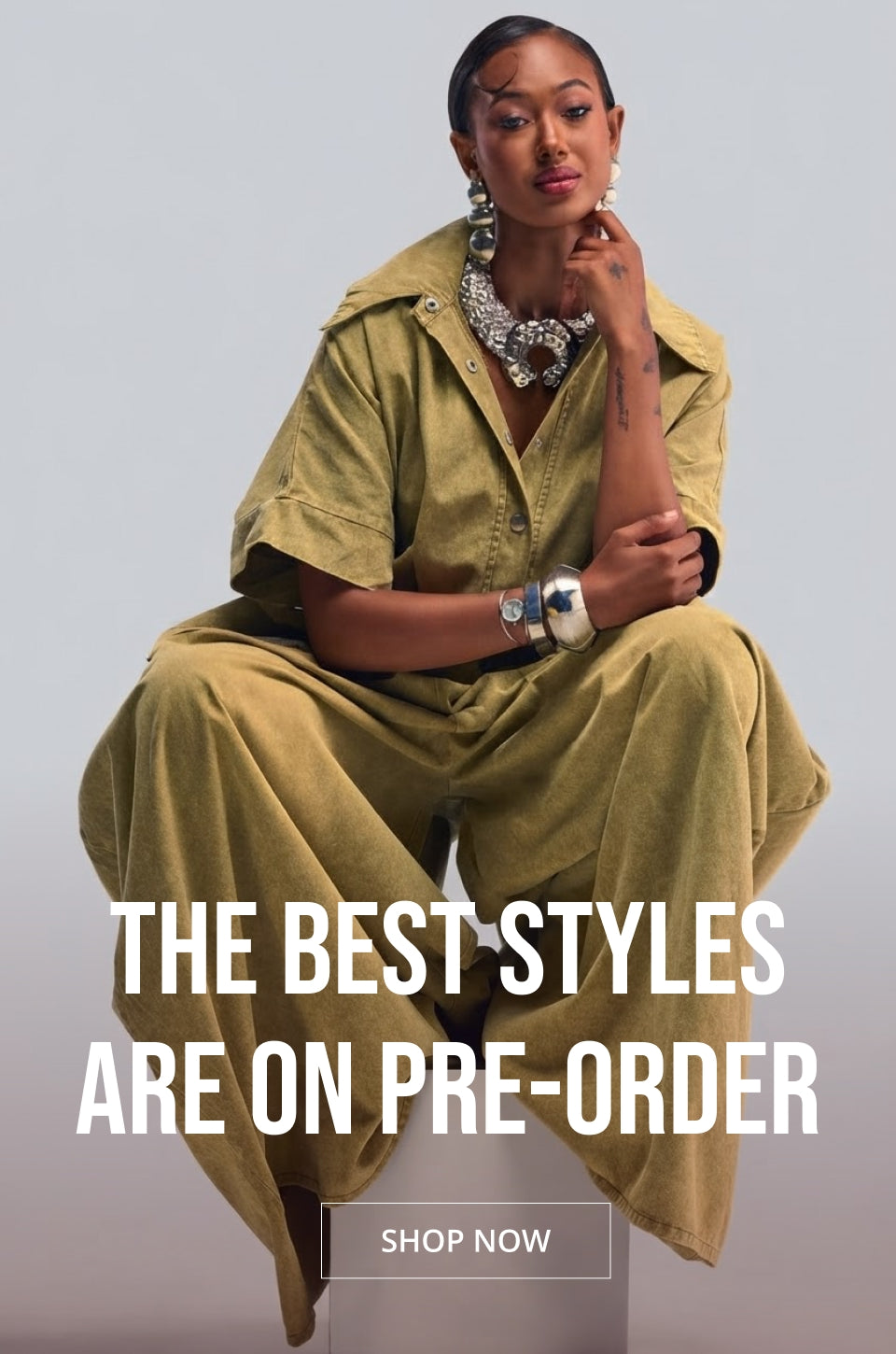 A model is seated against a light gray studio background wearing a matching olive green short-sleeve collared shirt and wide-leg trousers set, accessorized with a chunky silver necklace and bracelets. Large white text overlays read 'THE BEST STYLES ARE ON PRE-ORDER' with a 'SHOP NOW' button below.
