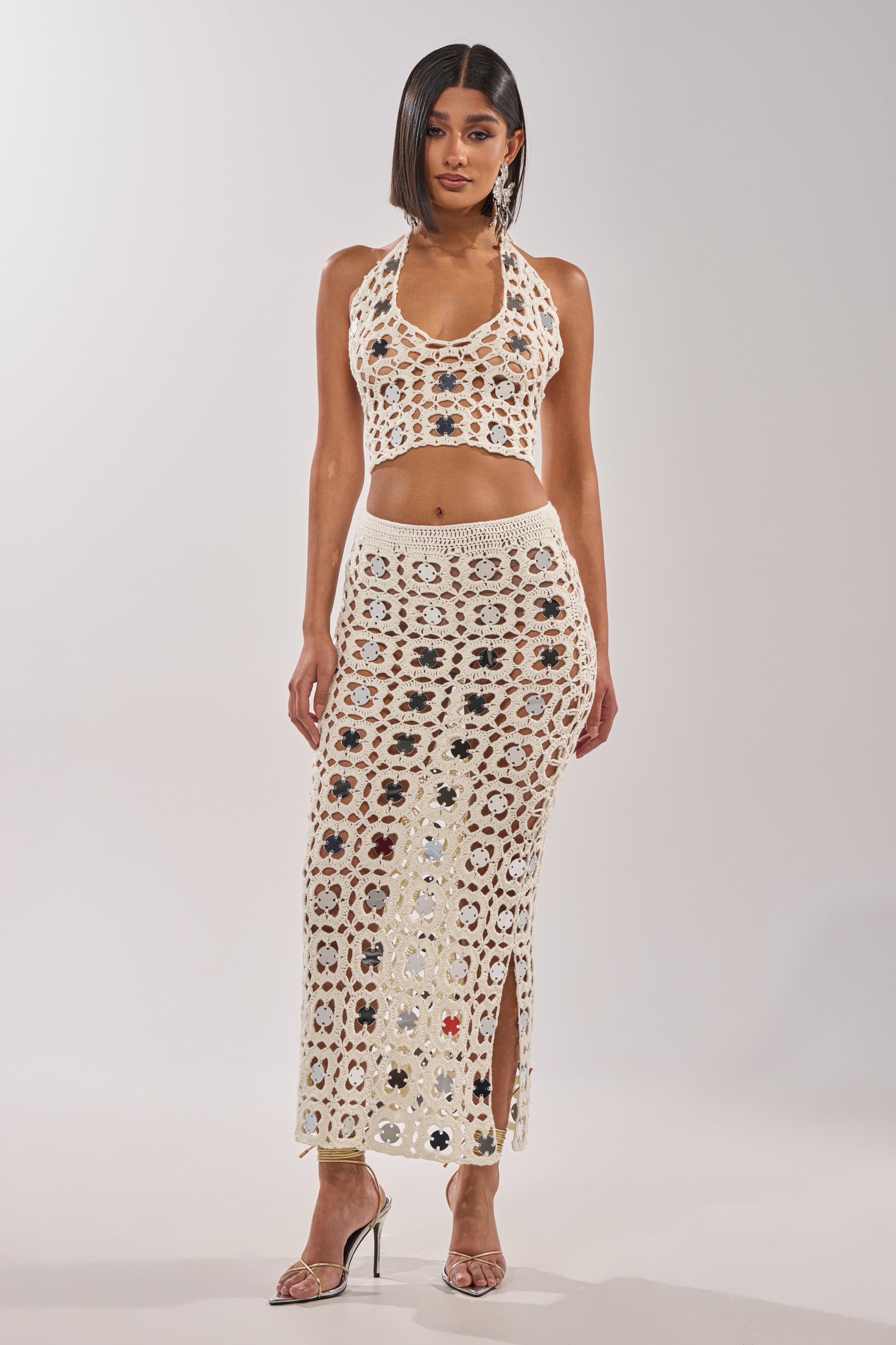 A woman wears the LETS GO TO THE RESORT MAXI SKIRT, a cream crochet maxi with a slit and multicolored florals, paired with a matching halter-neck crop top and heels for a chic resort look against a plain background. 0
