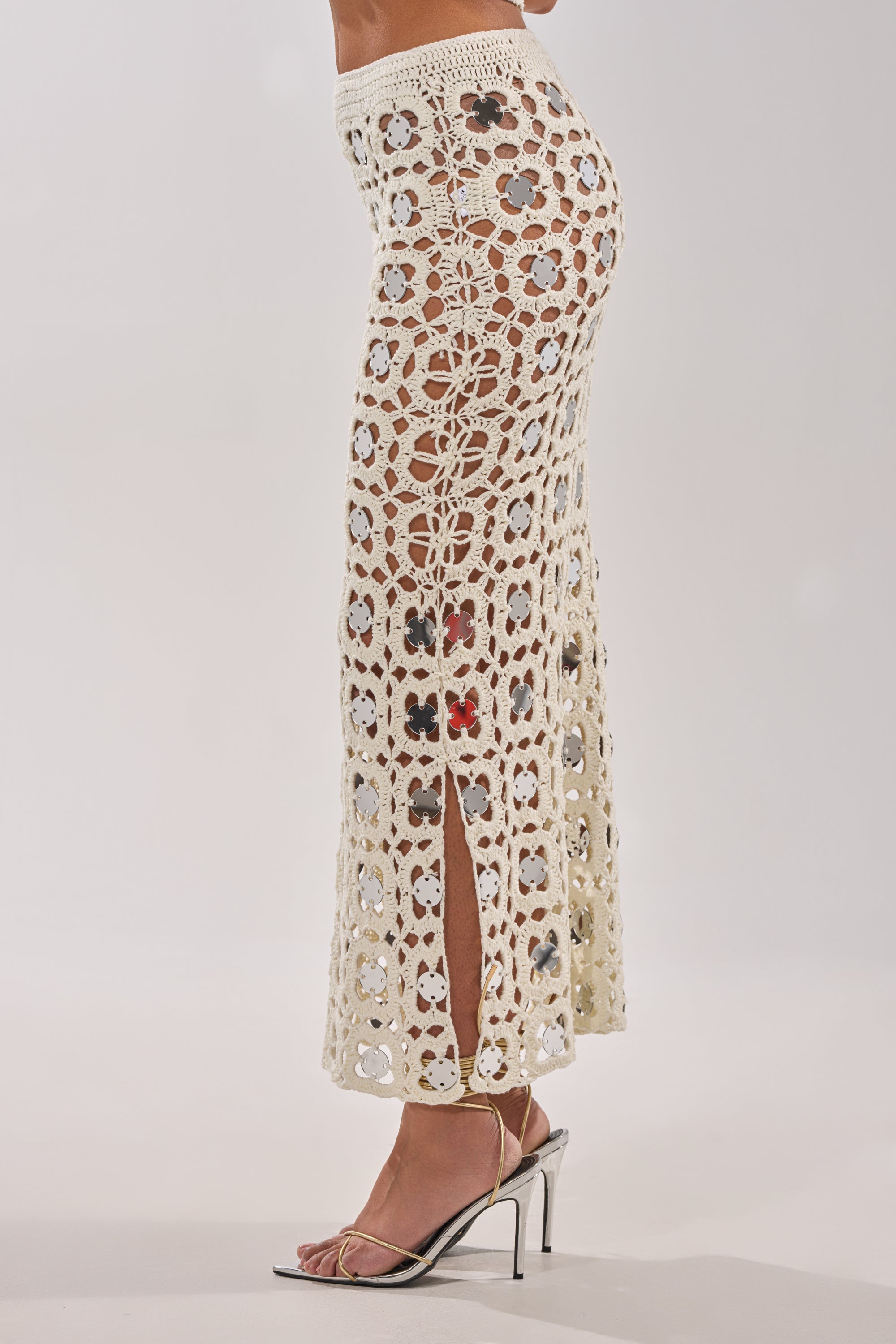 Wearing the LETS GO TO THE RESORT MAXI SKIRT—a cream crochet maxi with floral patterns, a side slit, and reflective discs—paired with metallic heels, this chic resort look stands out against a plain white background. 3