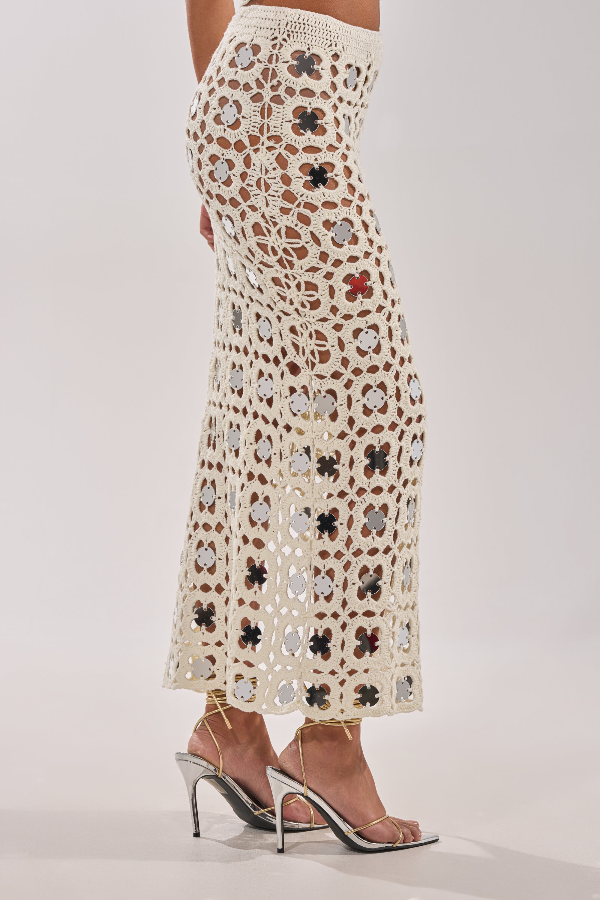 Wearing the LETS GO TO THE RESORT MAXI SKIRT—cream crochet with floral patterns and reflective discs—paired with metallic high-heeled sandals, this look channels effortless chic against a simple light backdrop. 4