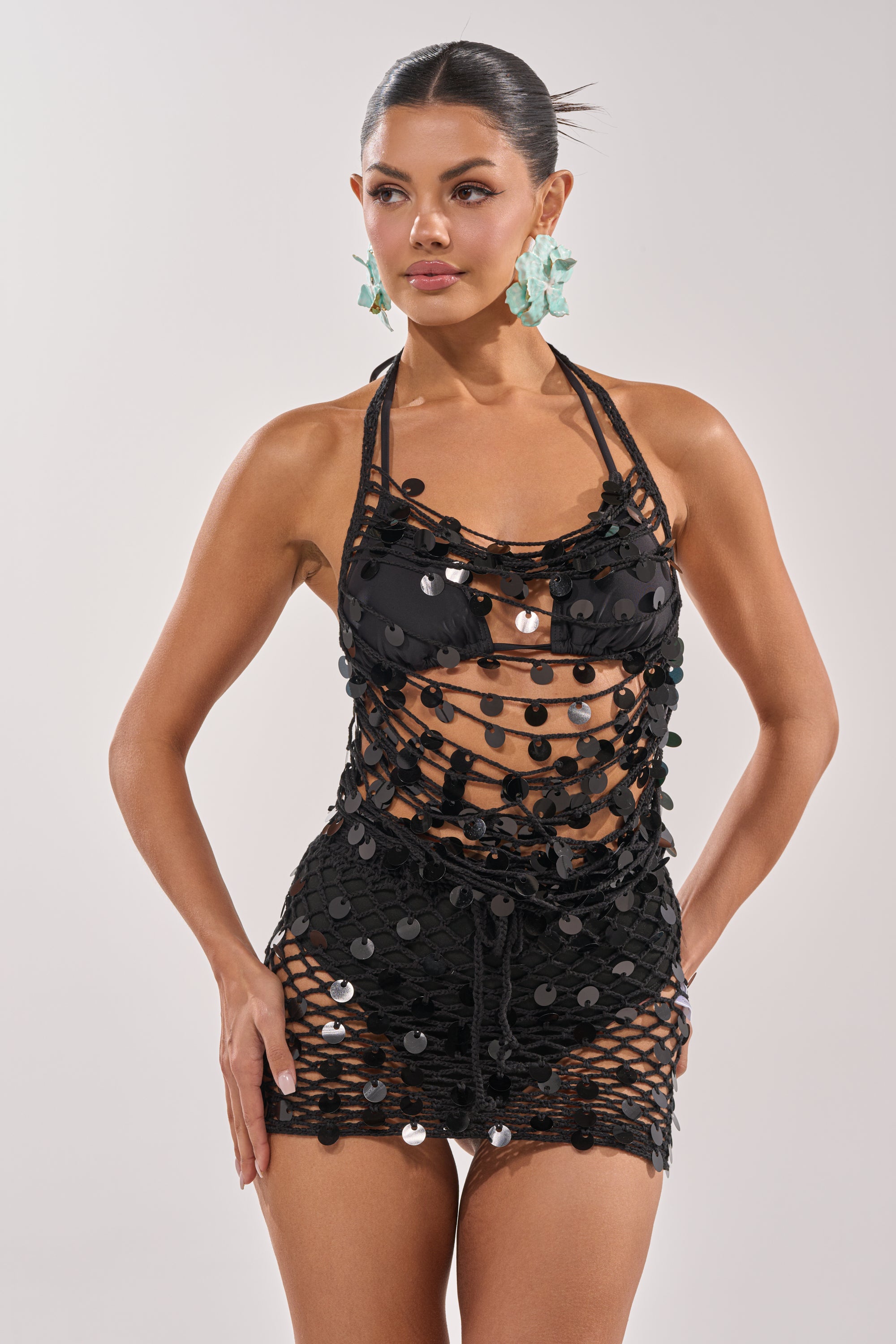A woman with slicked-back hair and large light blue earrings stands with one hand on her hip, wearing the DAYTIME VIBES CROCHET BLOUSE styled as a black, strappy, netted mini dress featuring a crochet cowl neck. 1