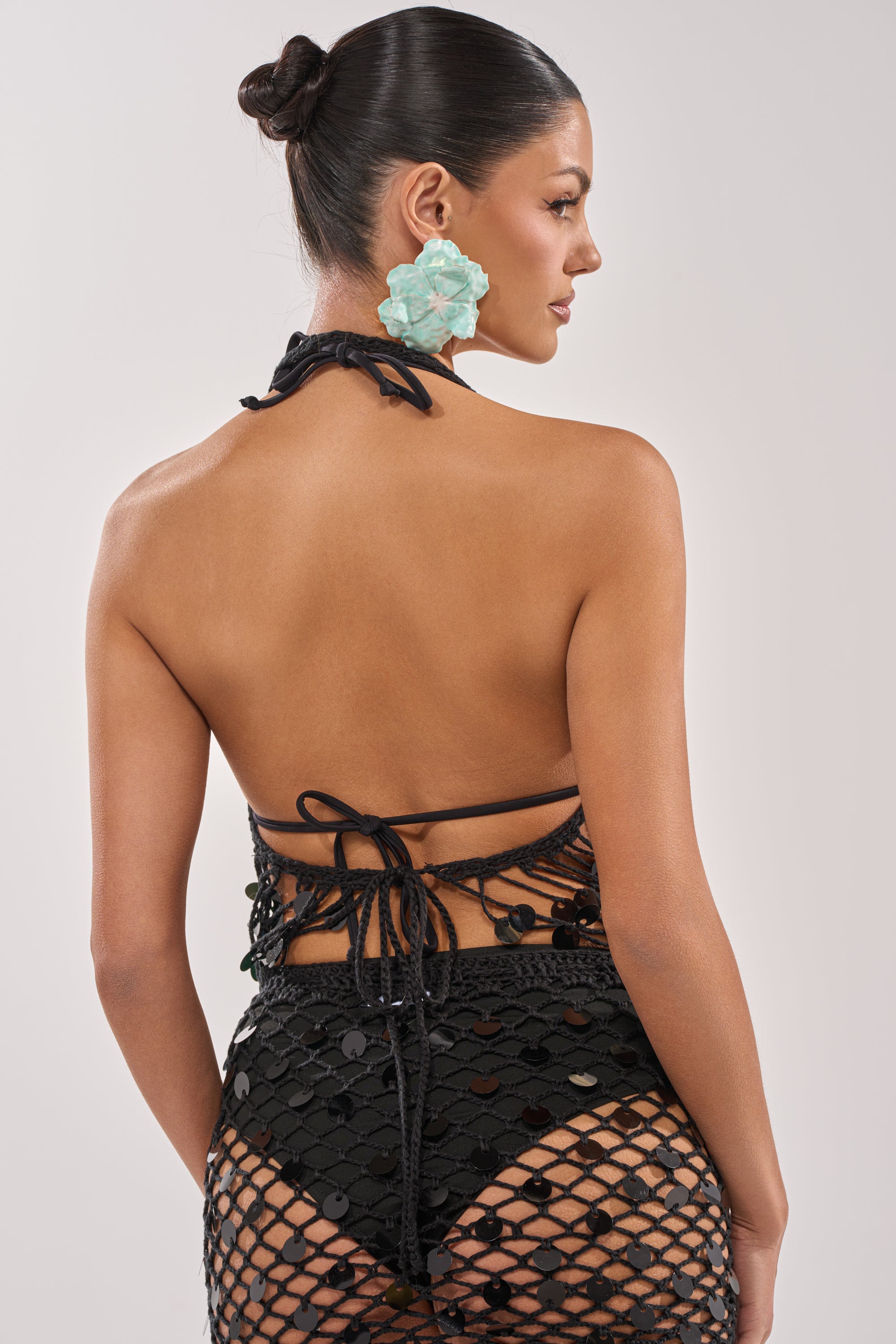 A woman with dark hair in a bun wears the DAYTIME VIBES CROCHET BLOUSE and large light blue flower-shaped earrings, standing against a plain light background. 3