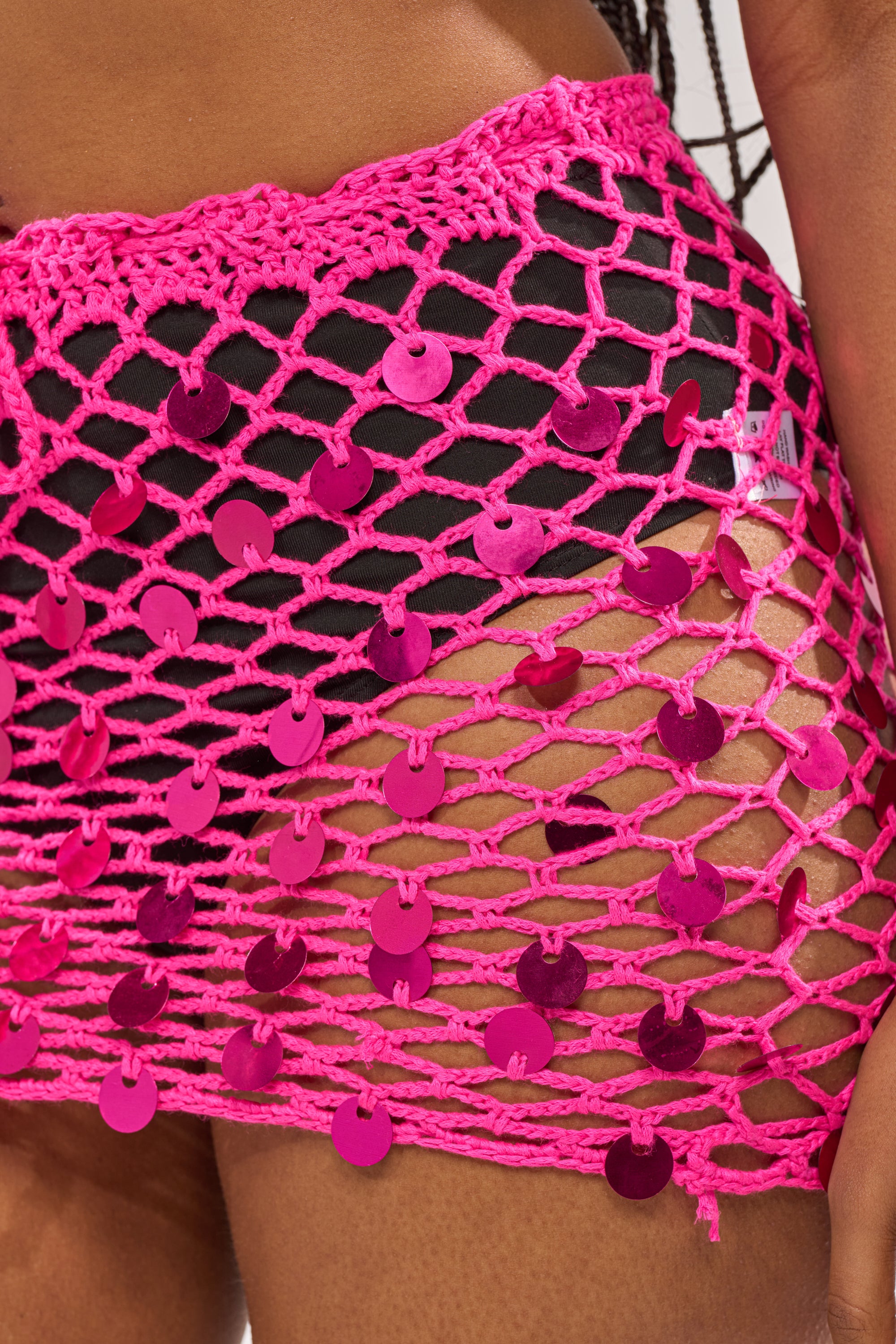 Close-up of a person wearing the HIGH VOLATGE MINI SKIRT, a hot pink crochet-knit high-rise mini skirt adorned with shiny pink sequins and an open pattern that reveals skin over black underwear. 8