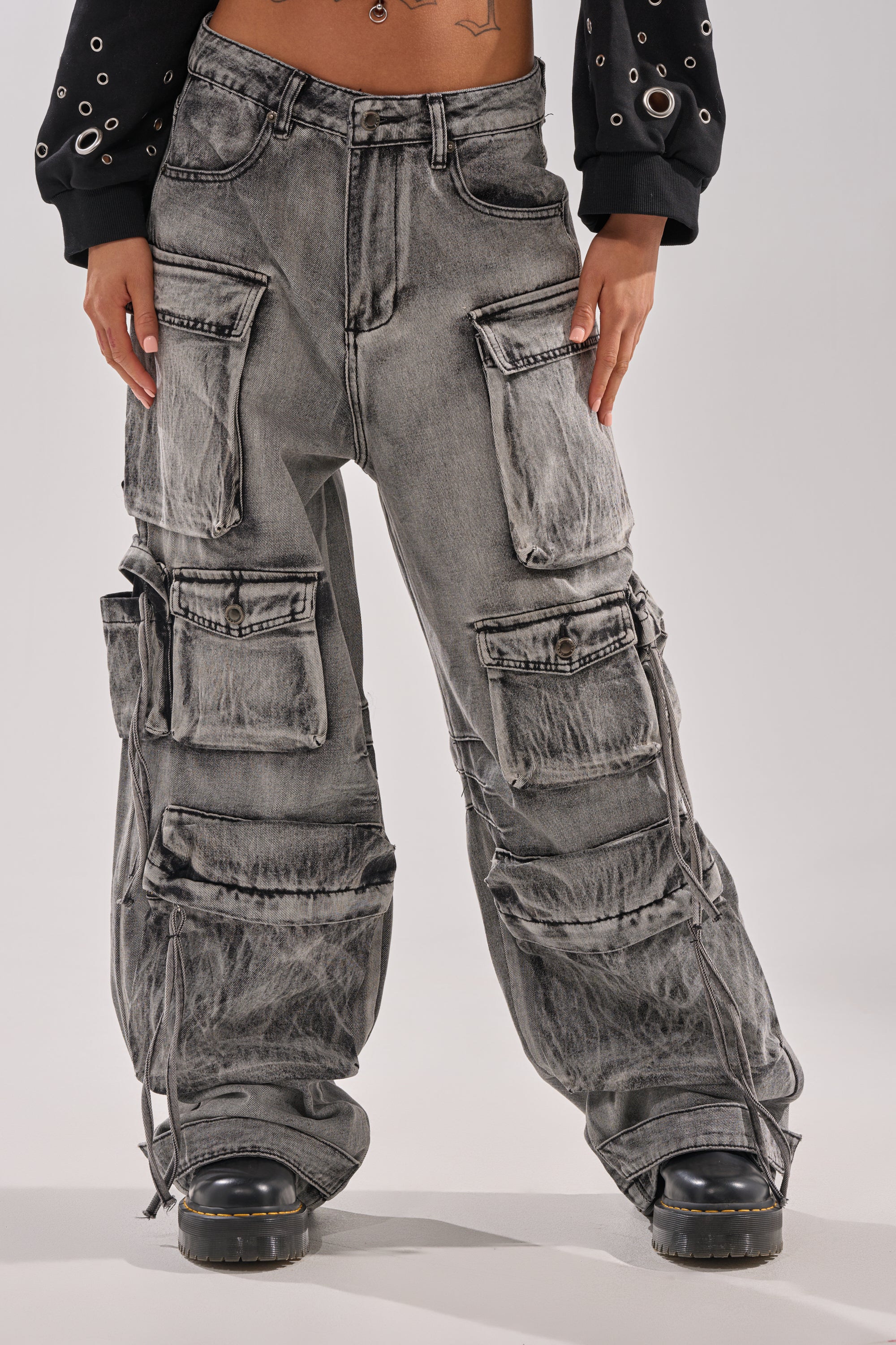 Wearing the CARGO FOR DAYS CARGO DENIM PANT with multiple large pockets, paired with black boots and a black long-sleeve top with metal eyelets; hands rest on thighs. 0