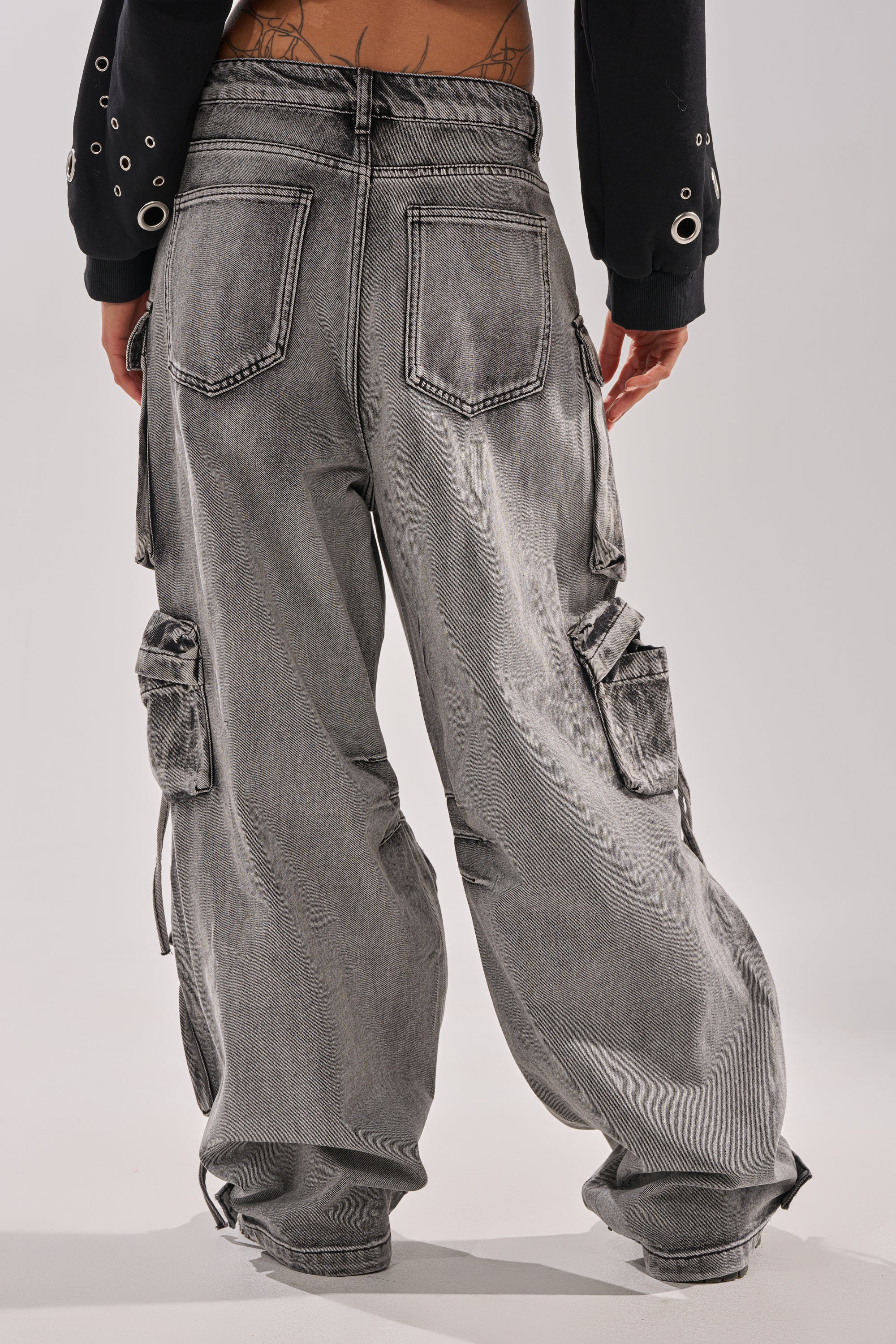 Model seen from the back wearing the CARGO FOR DAYS CARGO DENIM PANT in loose, light gray acid-wash style with multiple cargo pockets and a low-rise waistband, partially revealing a lower back tattoo. 3