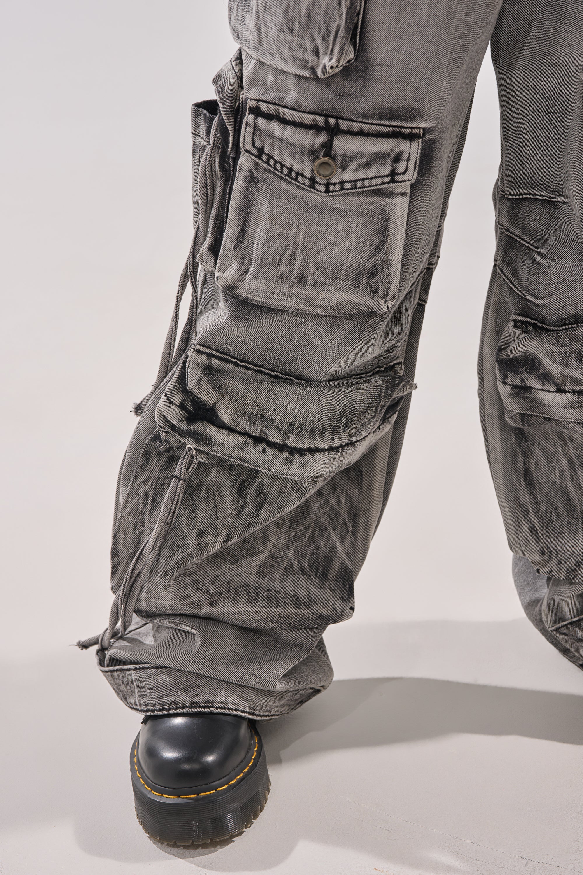 A close-up of a person wearing the CARGO FOR DAYS CARGO DENIM PANT—wide-leg jeans with multiple large pockets and drawstrings—paired with chunky black platform shoes against a plain background. 6