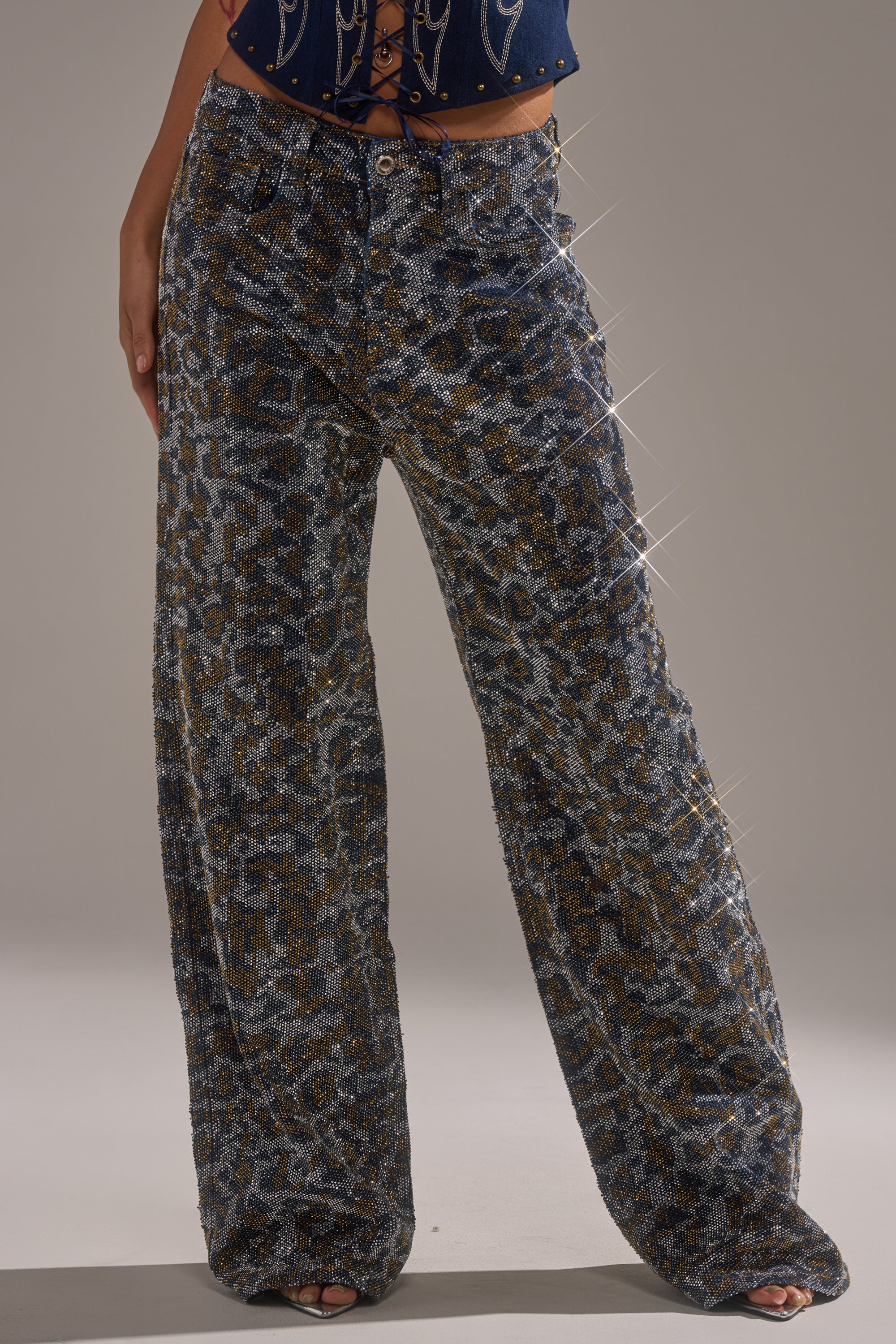 A person wearing the CHEETAH GIRLY RHINESTONE DENIM PANT—high-waisted, wide-leg jeans featuring a metallic leopard print—stands with one hand at their side; only their lower torso and legs are visible. 0