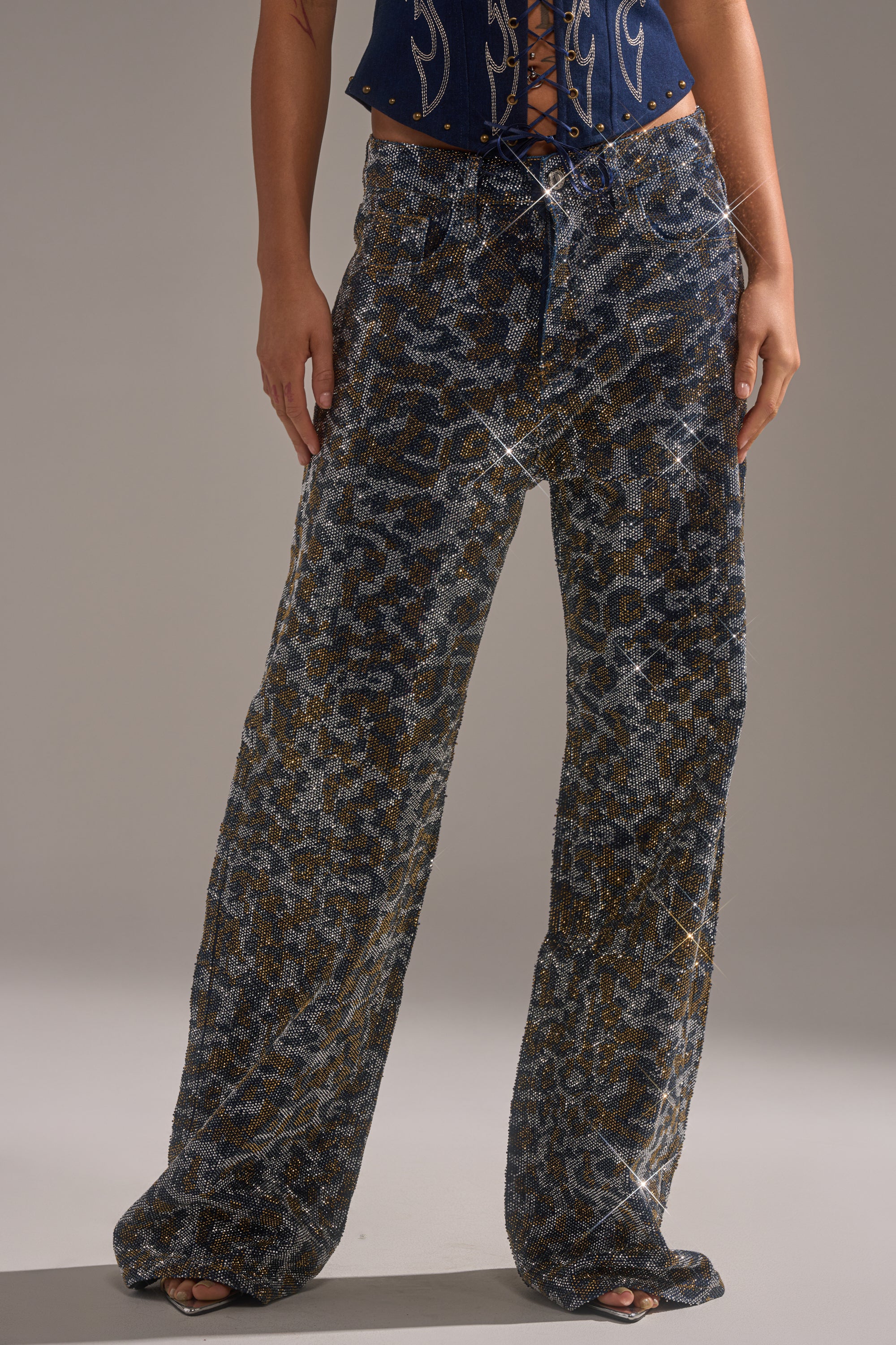 A person models the CHEETAH GIRLY RHINESTONE DENIM PANT—sparkly, wide-leg jeans with a silver and gold animal print—paired with a navy blue corset top. The image highlights the pants from waist to feet. 1